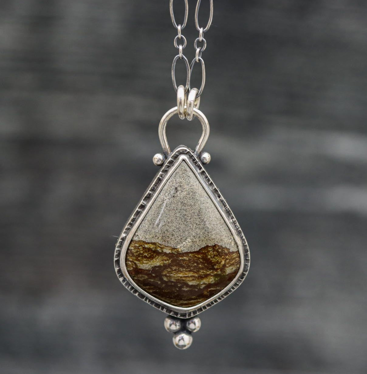 A sterling silver pendant featuring a Disaster Peak picture jasper gemstone, with a textured metal surface, mounted on an oval link chain.