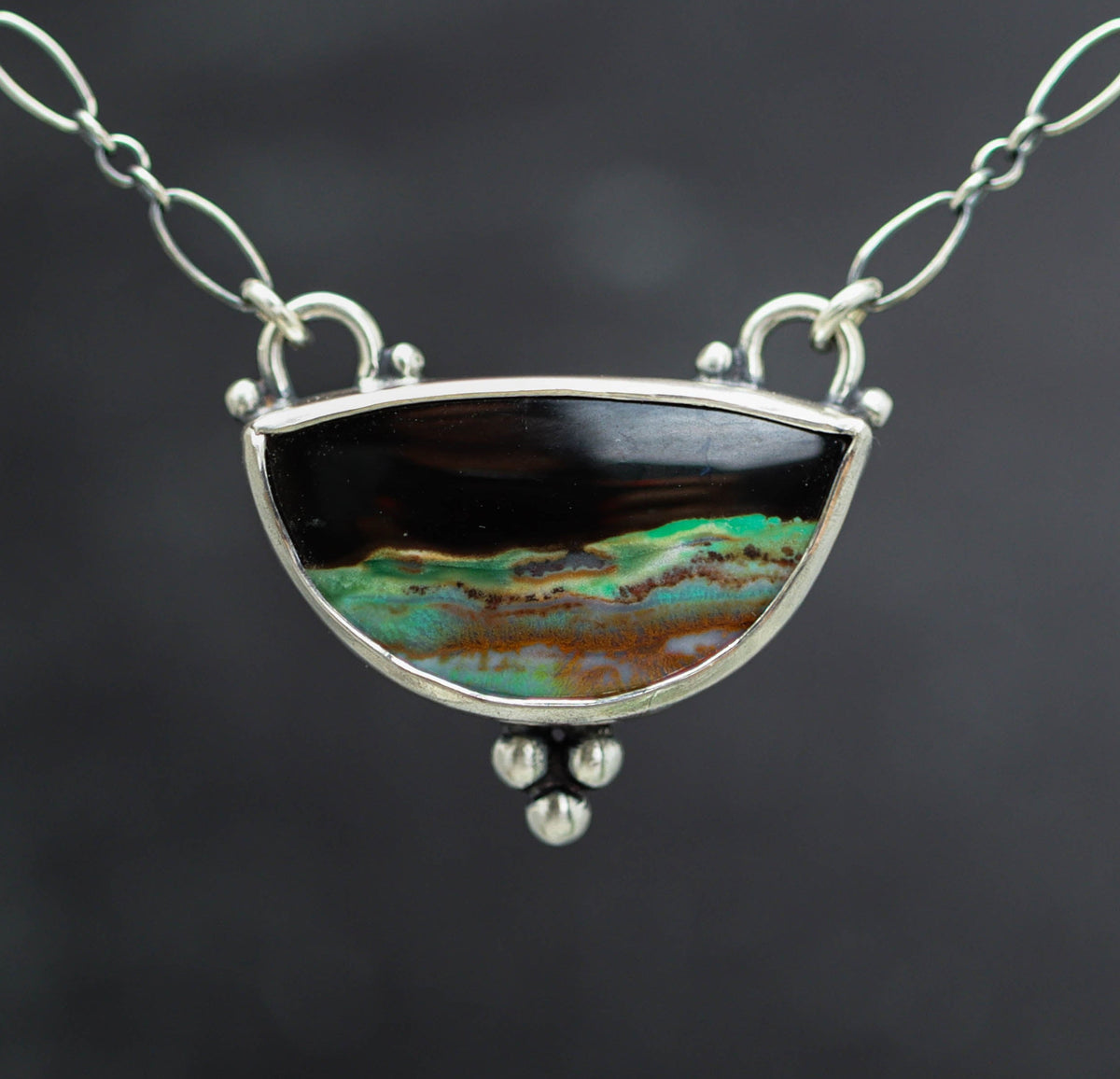 A silver link chain necklace featuring a semi-circular pendant with a black top section and a bottom section displaying a vibrant green, turquoise, and brown marbled pattern, set against a dark gray background.