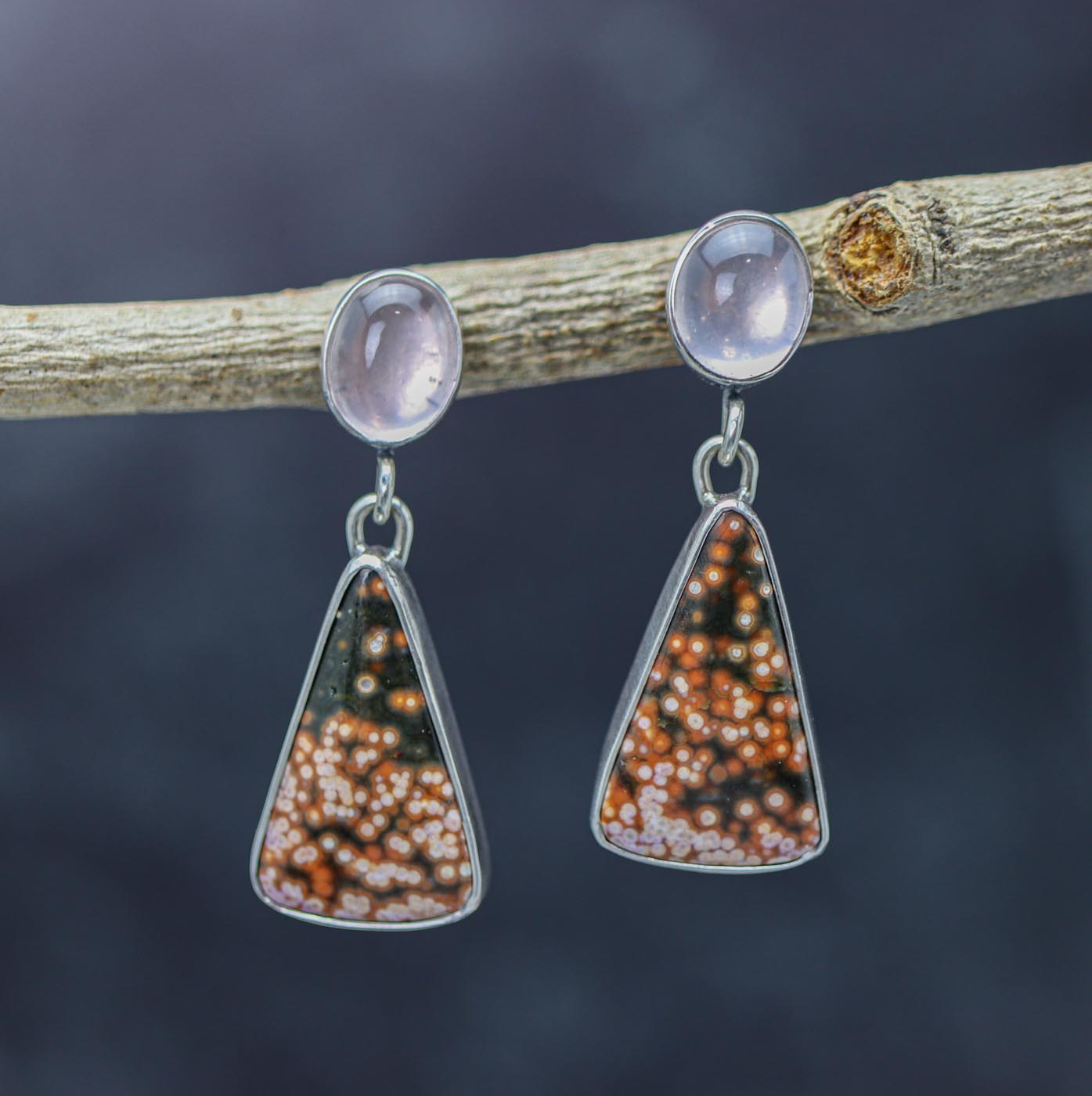 A pair of sterling silver dangle earrings featuring ocean jasper and rose quartz stones, displayed against a natural outdoor background.