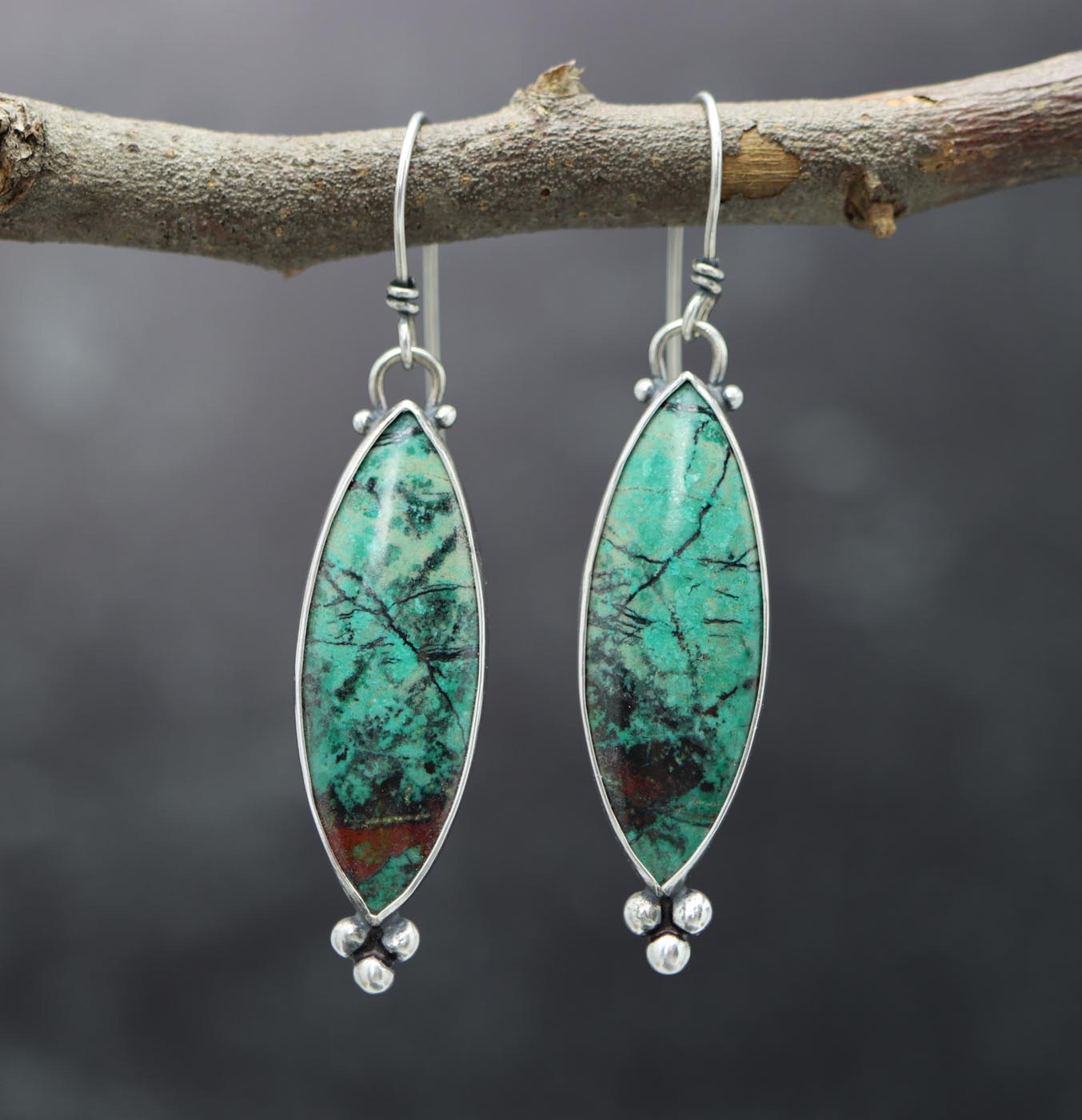 A pair of sterling silver dangle earrings with green sonoran sunrise stones, red cuprite, and black matrix accents, displayed on a branch.