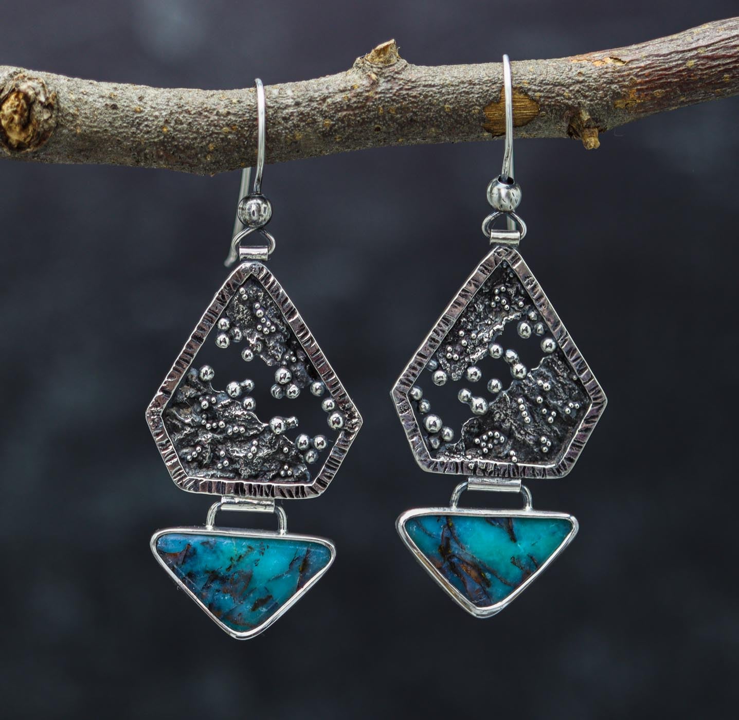 A pair of silver earrings with reticulated and granulated designs, featuring petrified opal wood inlays, displayed against a natural outdoor background.