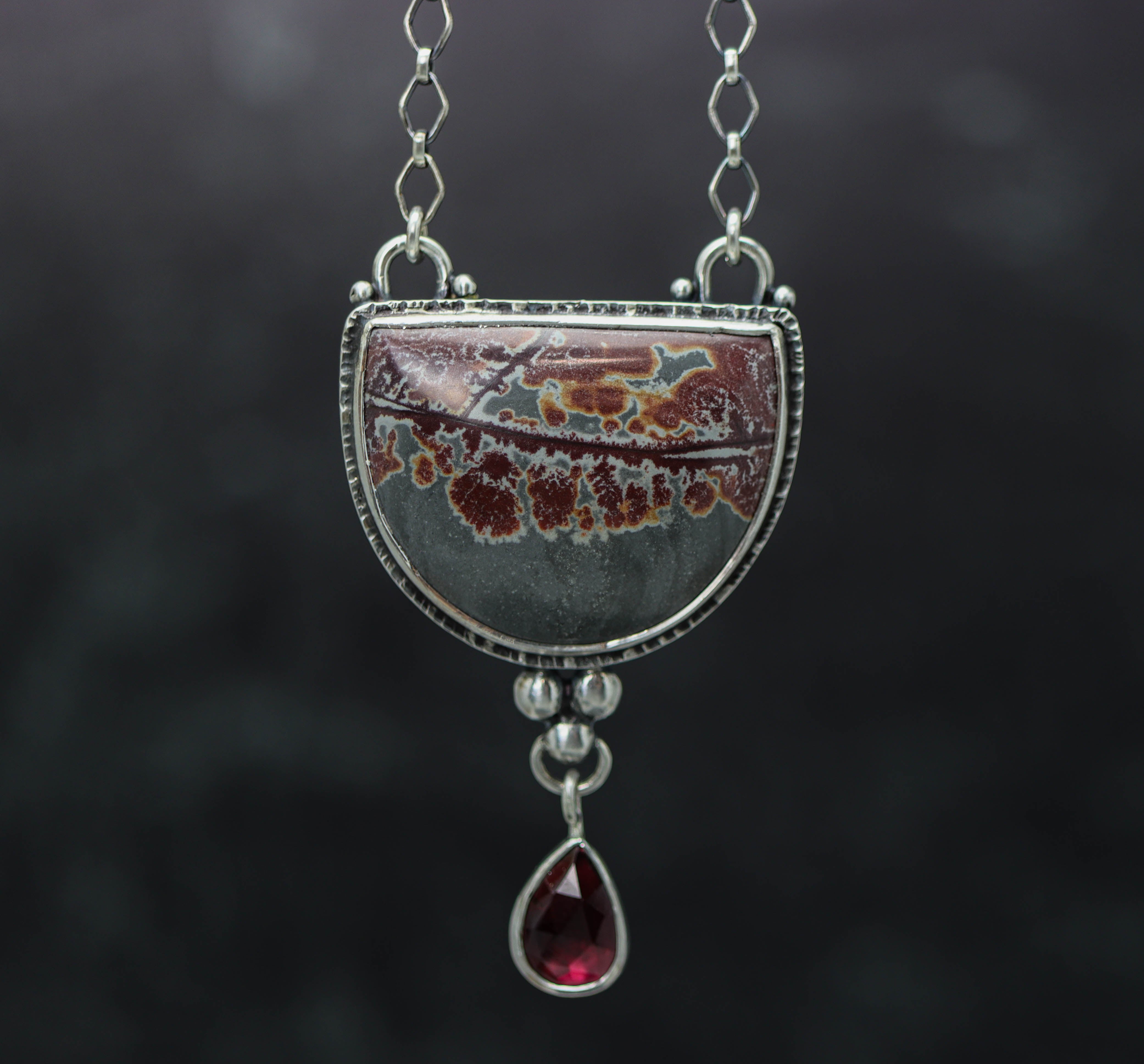 A sterling silver pendant featuring a sonoran dendritic rhyolite jasper gemstone with red dendrites, and a drop of garnet, attached to a sterling silver chain.