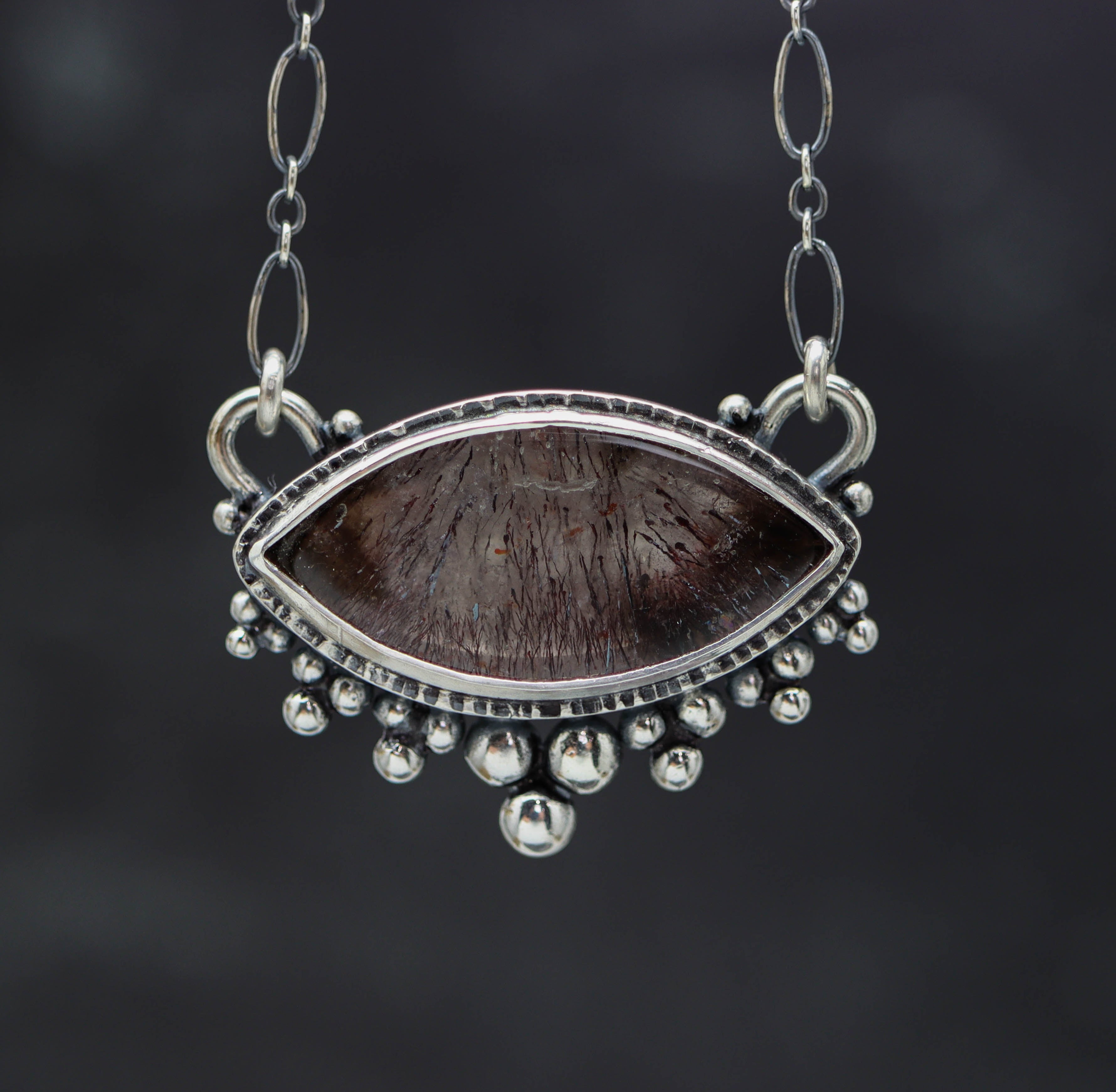 A sterling silver pendant featuring a super seven or melody stone gemstone, with a double bezel and ruffle of silver balls, on an oval link chain.