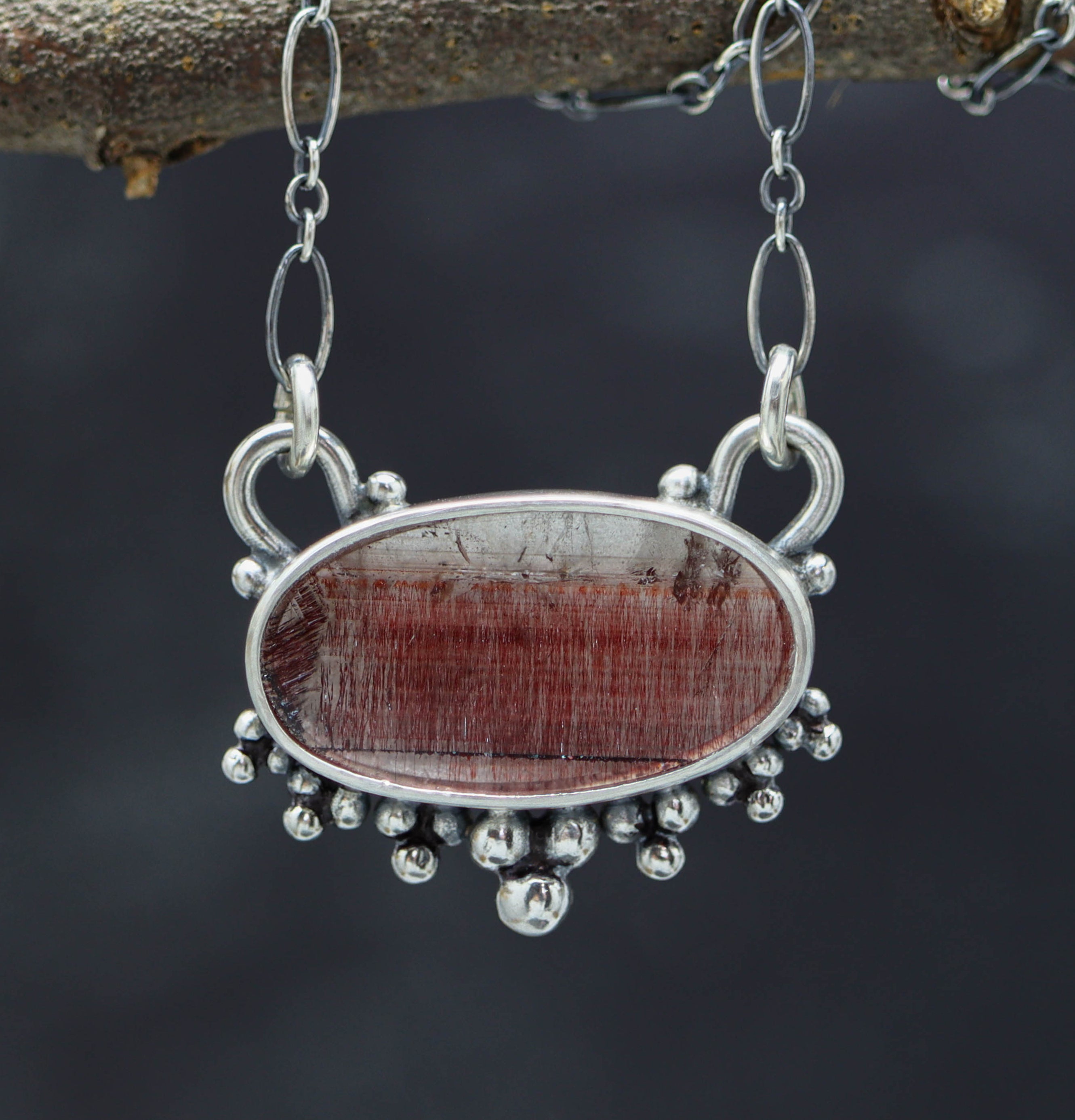 A sterling silver pendant featuring a red gemstone, possibly a Super Seven or Melody Stone, set in a double bezel design with silver balls textured on the back. The pendant is suspended from an oval link chain with a lobster claw closure.