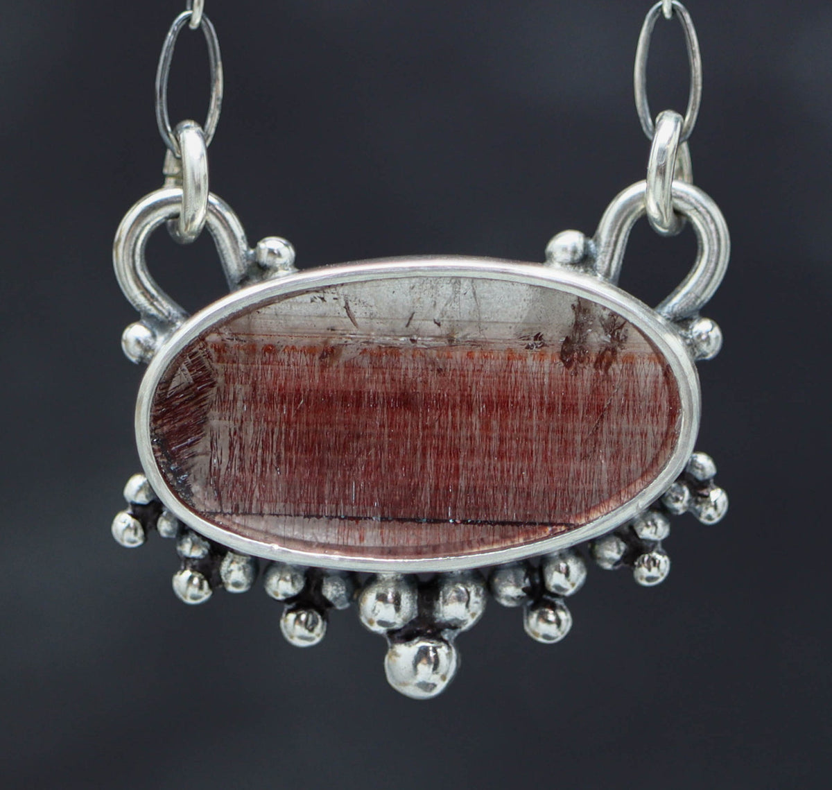 A sterling silver pendant featuring a red gemstone, possibly a Super Seven or Melody Stone, set in a double bezel design with silver balls textured on the back. The pendant is suspended from an oval link chain with a lobster claw closure.