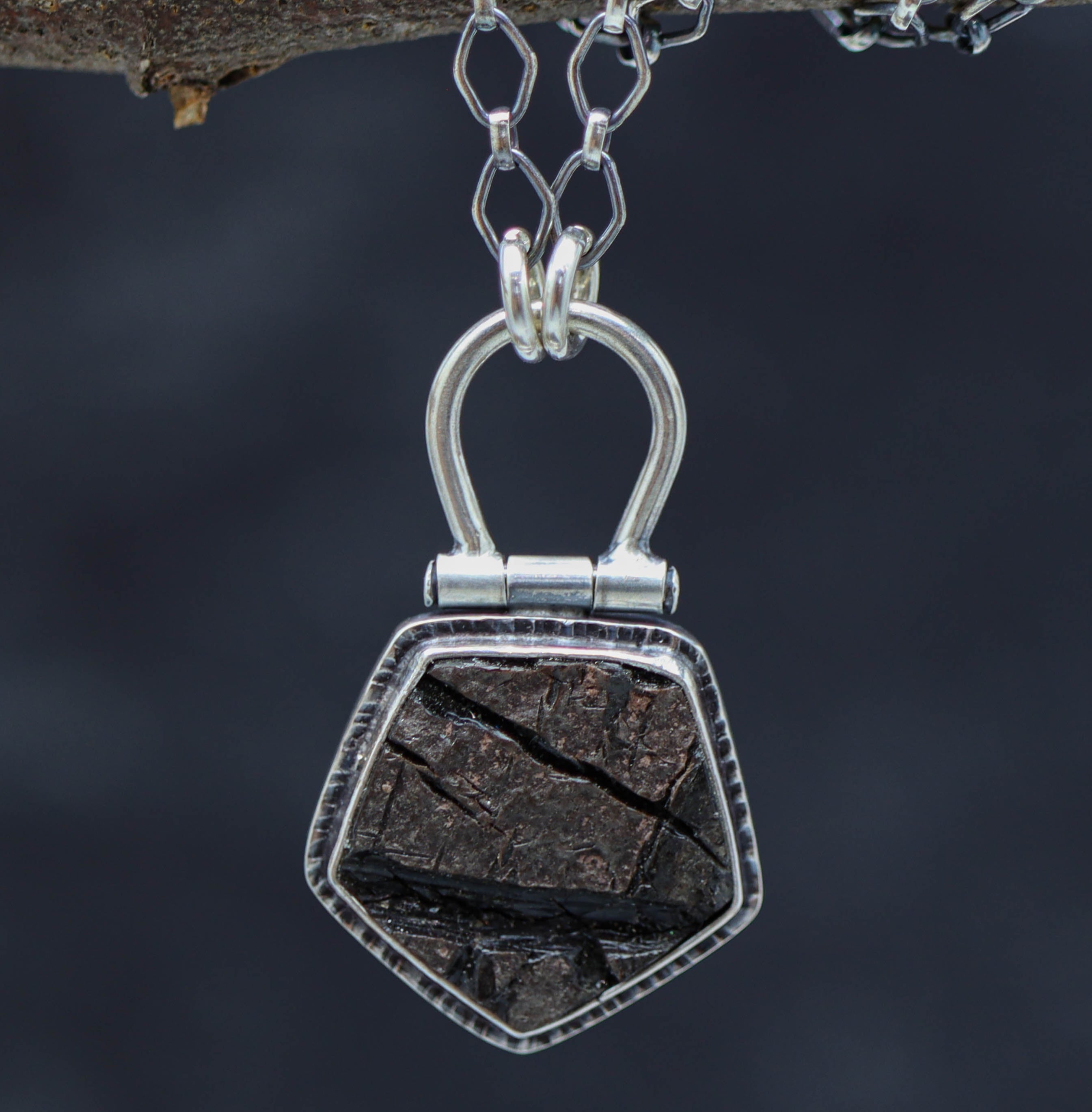 A sterling silver necklace with a natural surface obsidian pendant, displayed on a tree branch.