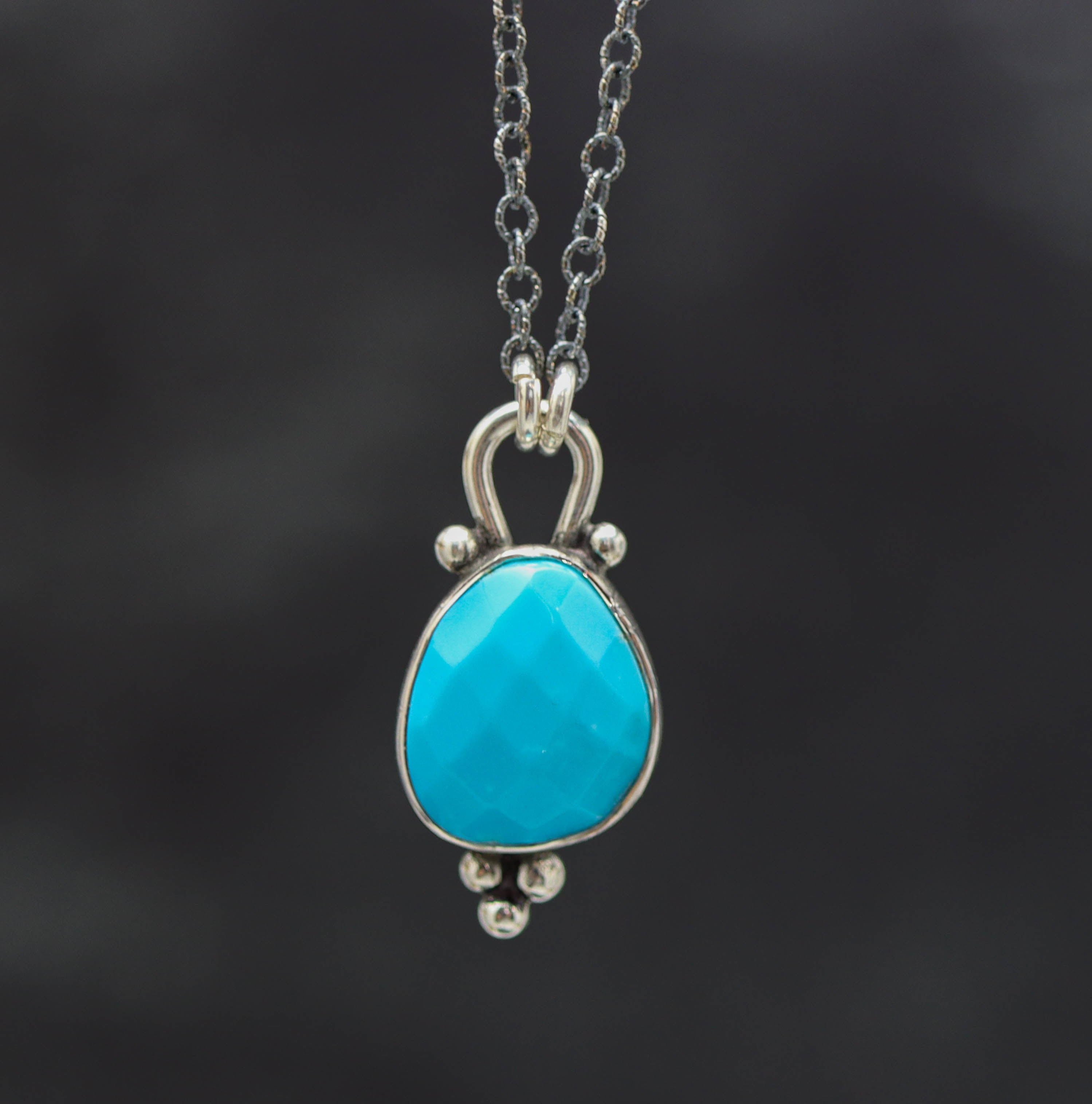A sterling silver pendant necklace with a faceted bright blue turquoise centerpiece, surrounded by silver ball accents on a dark background.