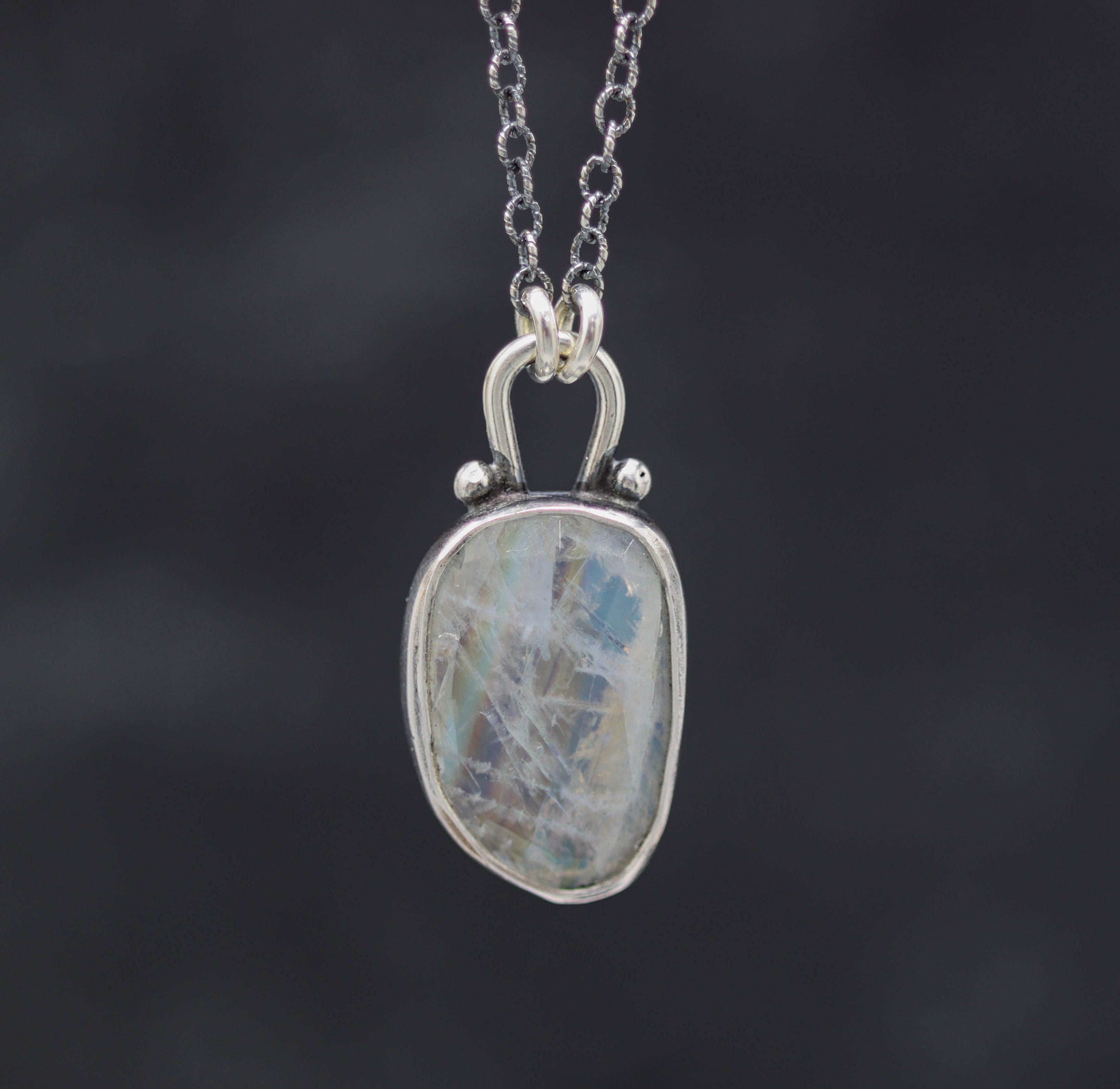 A sterling silver pendant necklace with a rainbow moonstone centerpiece and small silver ball accents, displayed on a silver chain.