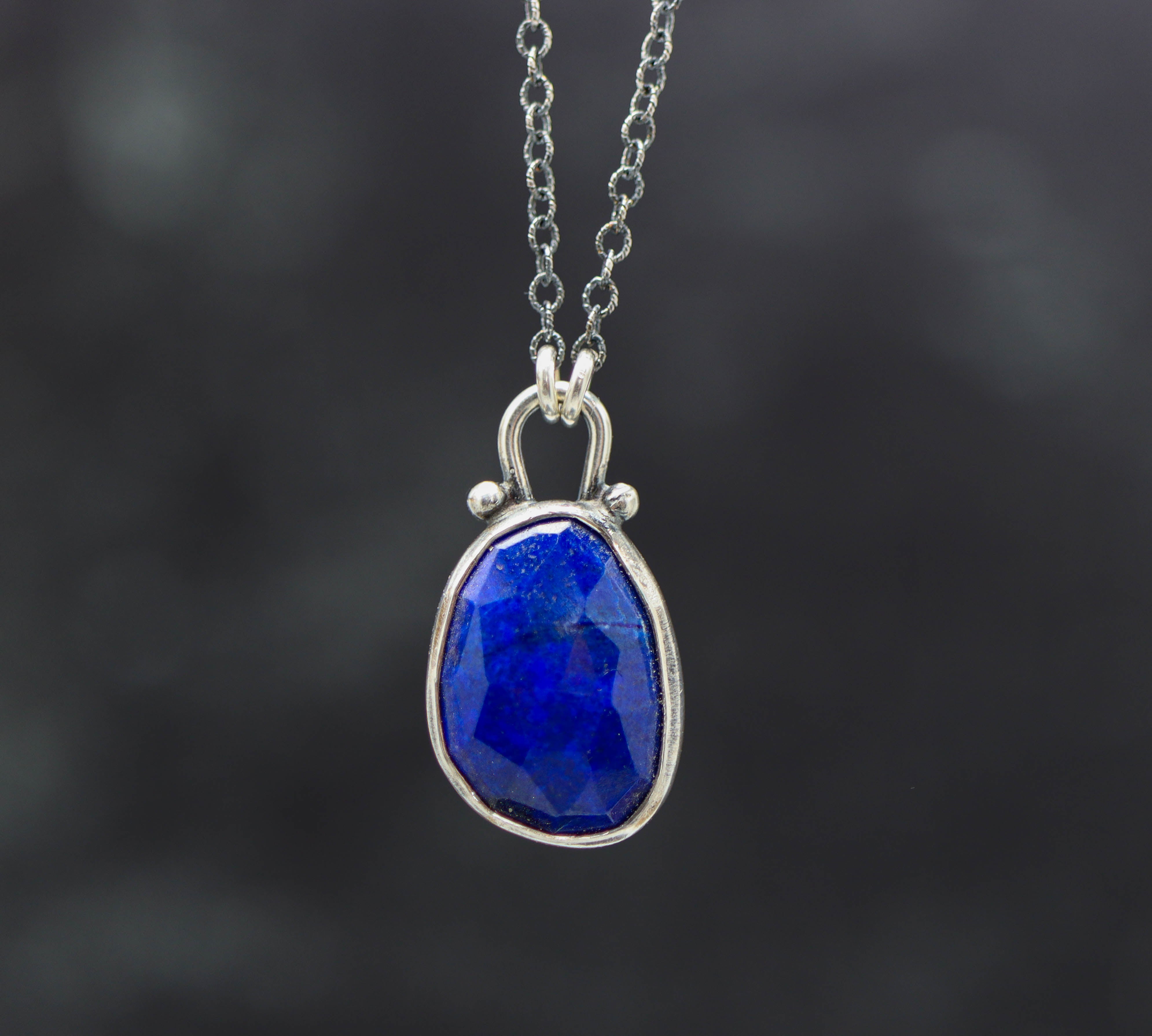 A sterling silver pendant necklace featuring a faceted rose cut blue lapis gemstone, with small silver ball accents, displayed on a silver cable link chain.