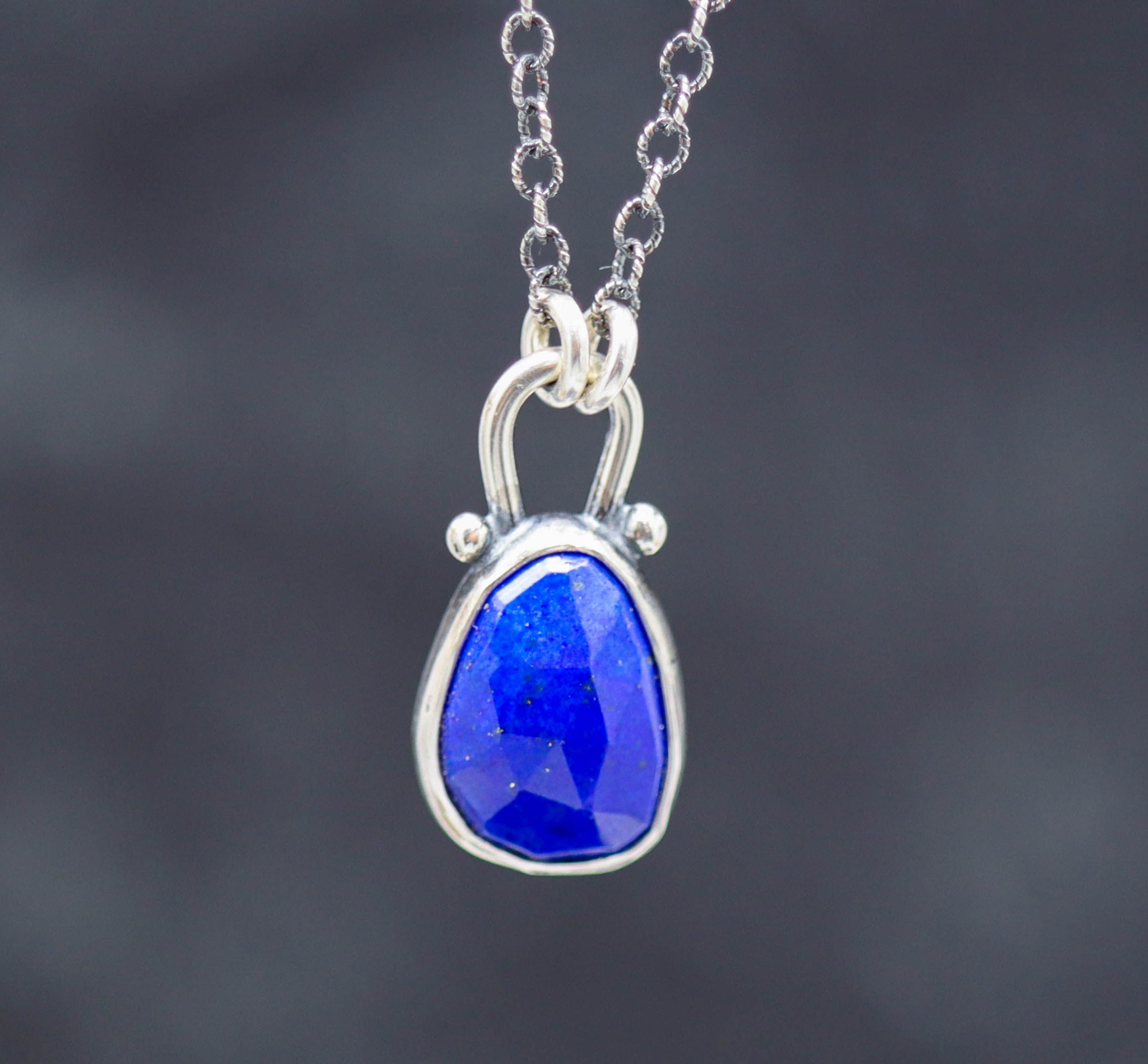 A sterling silver pendant necklace featuring a faceted rose cut blue lapis centerpiece with small silver ball accents, suspended from an oxidized cable link chain.