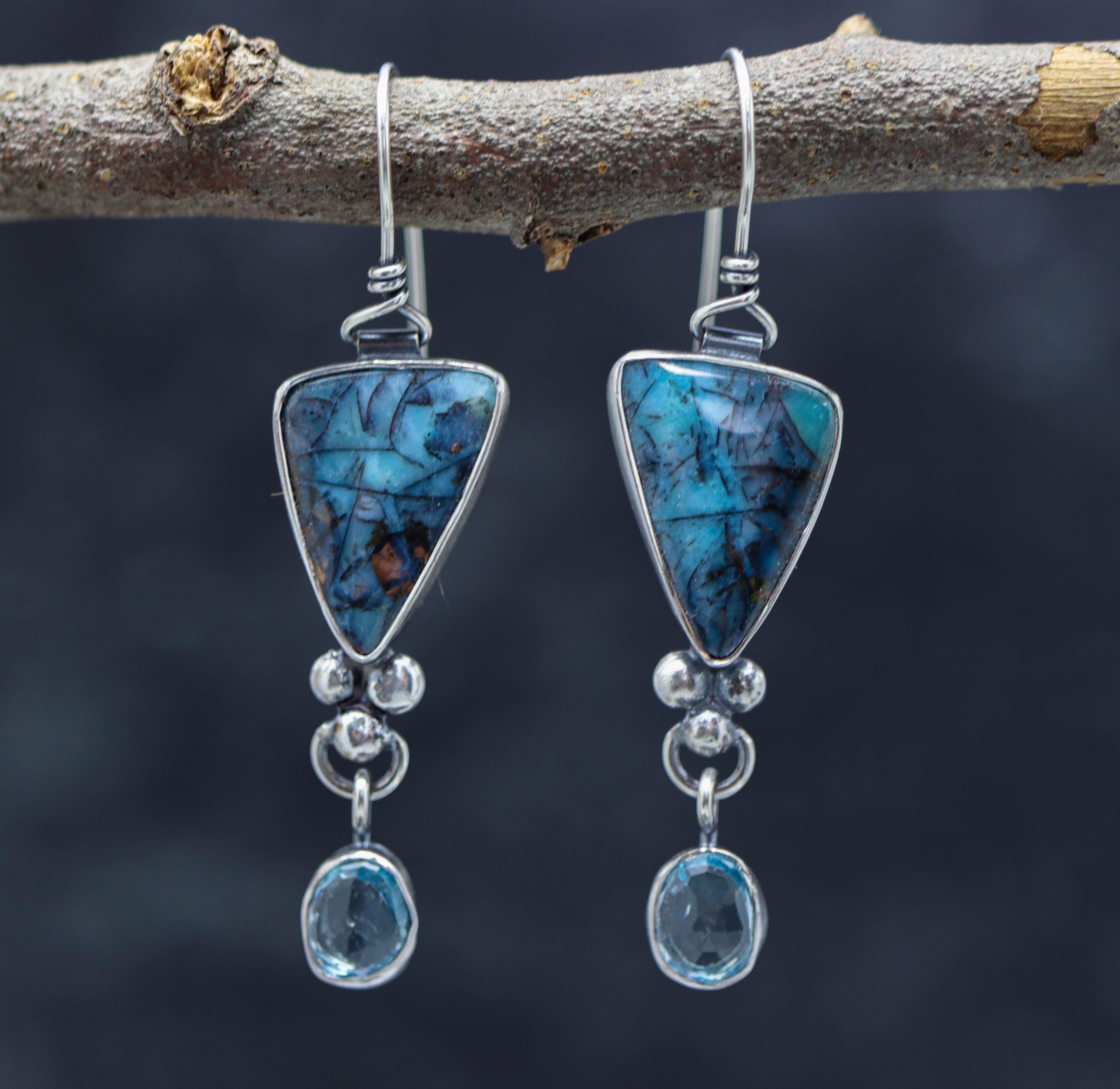 A pair of sterling silver dangle earrings featuring an opalized petrified wood design with blue topaz, displayed against a natural outdoor background.
