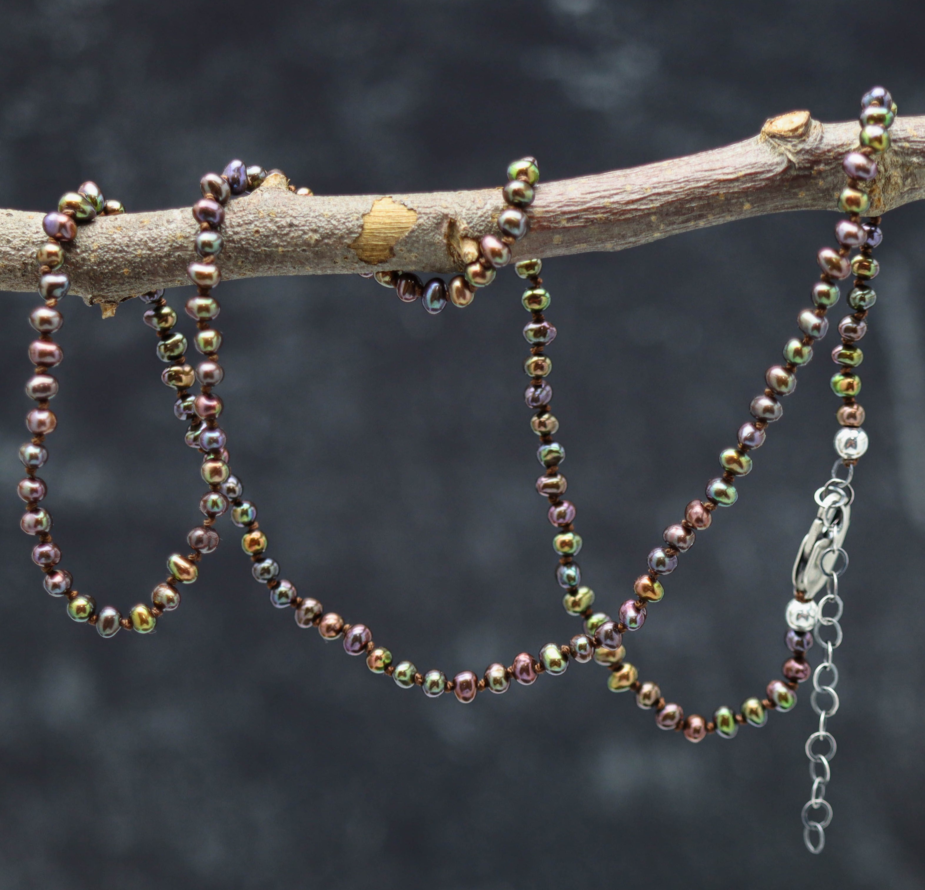 A hand knotted necklace made of peacock bronze freshwater pearls, displayed on a branch, with sterling silver closure.