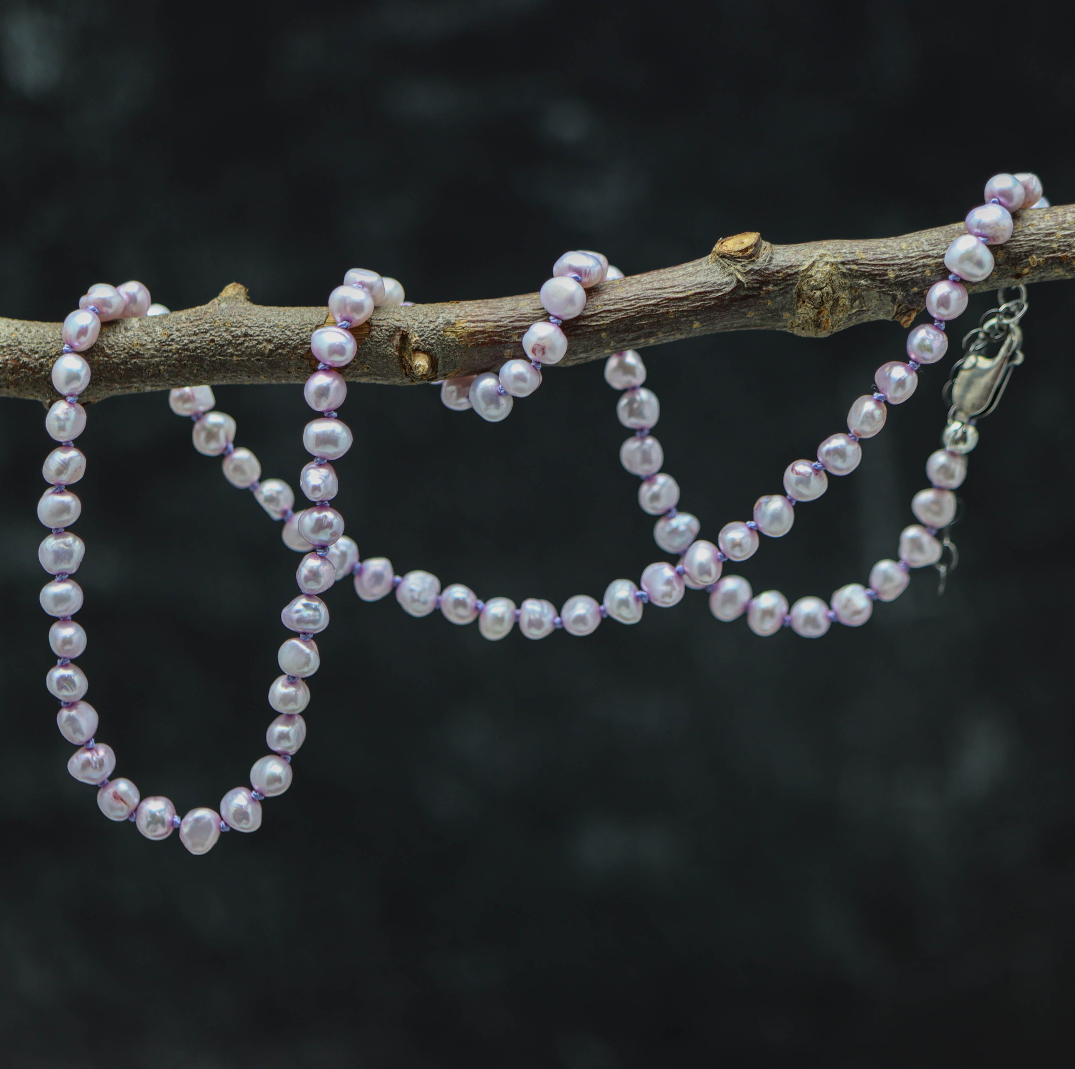 A hand knotted necklace made of lavender freshwater pearls, strung on silk thread with a sterling silver clasp, displayed on a branch against a dark background.
