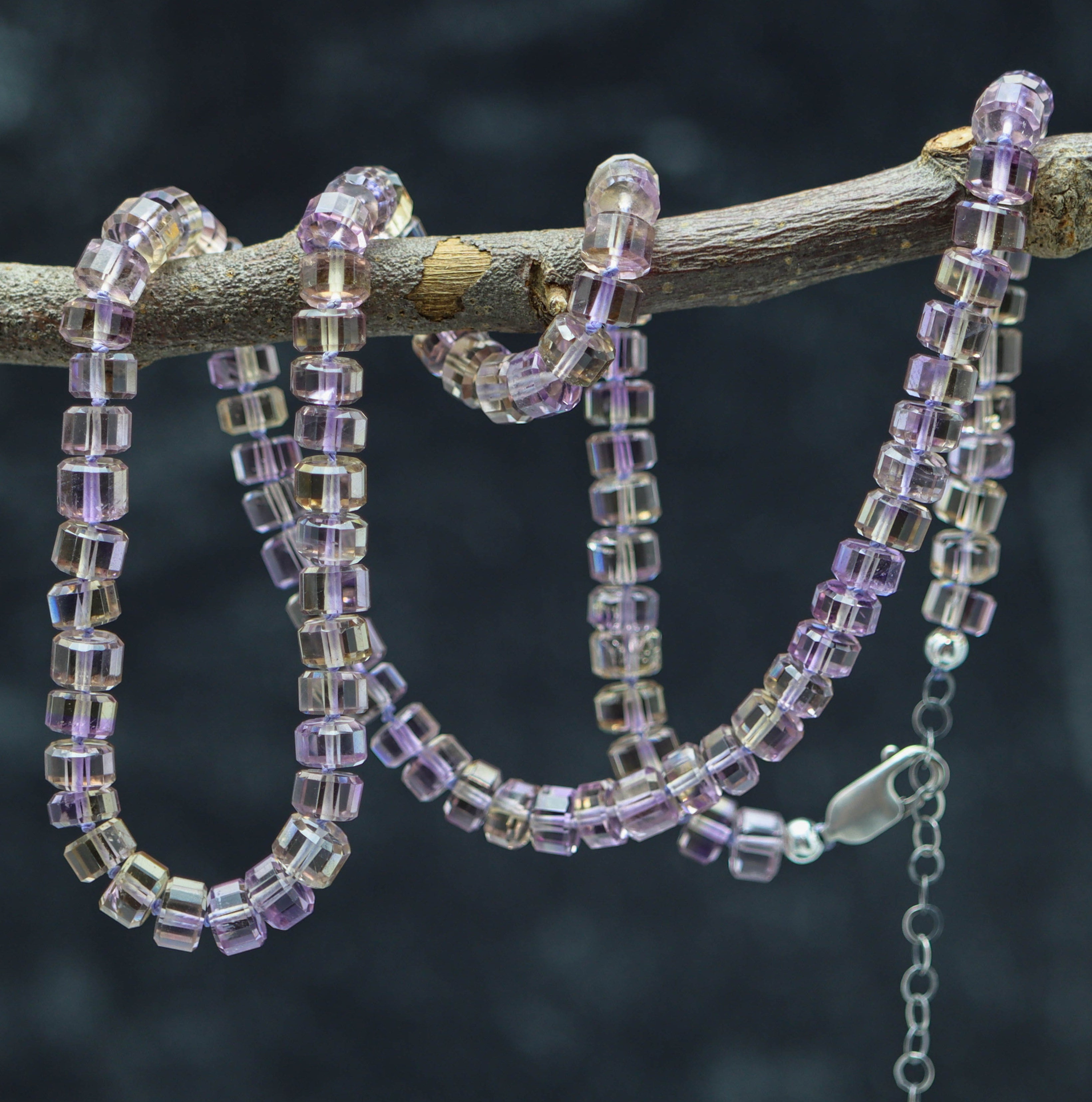 A hand knotted bead necklace with sterling silver faceted barrels, featuring a blend of purple and clear beads, displayed on a branch against a dark background.