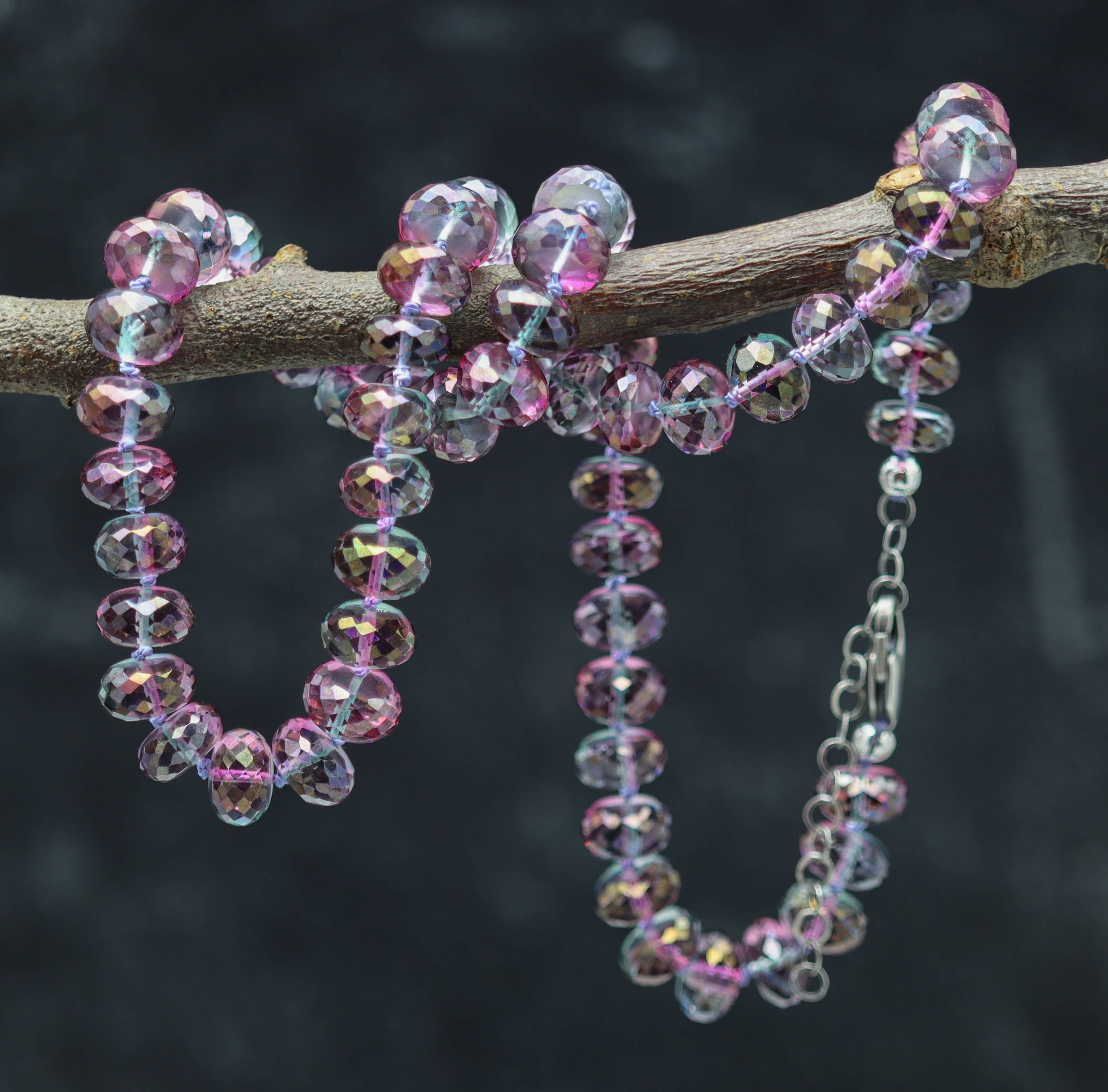 A hand knotted bead necklace made of multicolored (purple, pink, blue) beads and sterling silver, displayed on a branch.