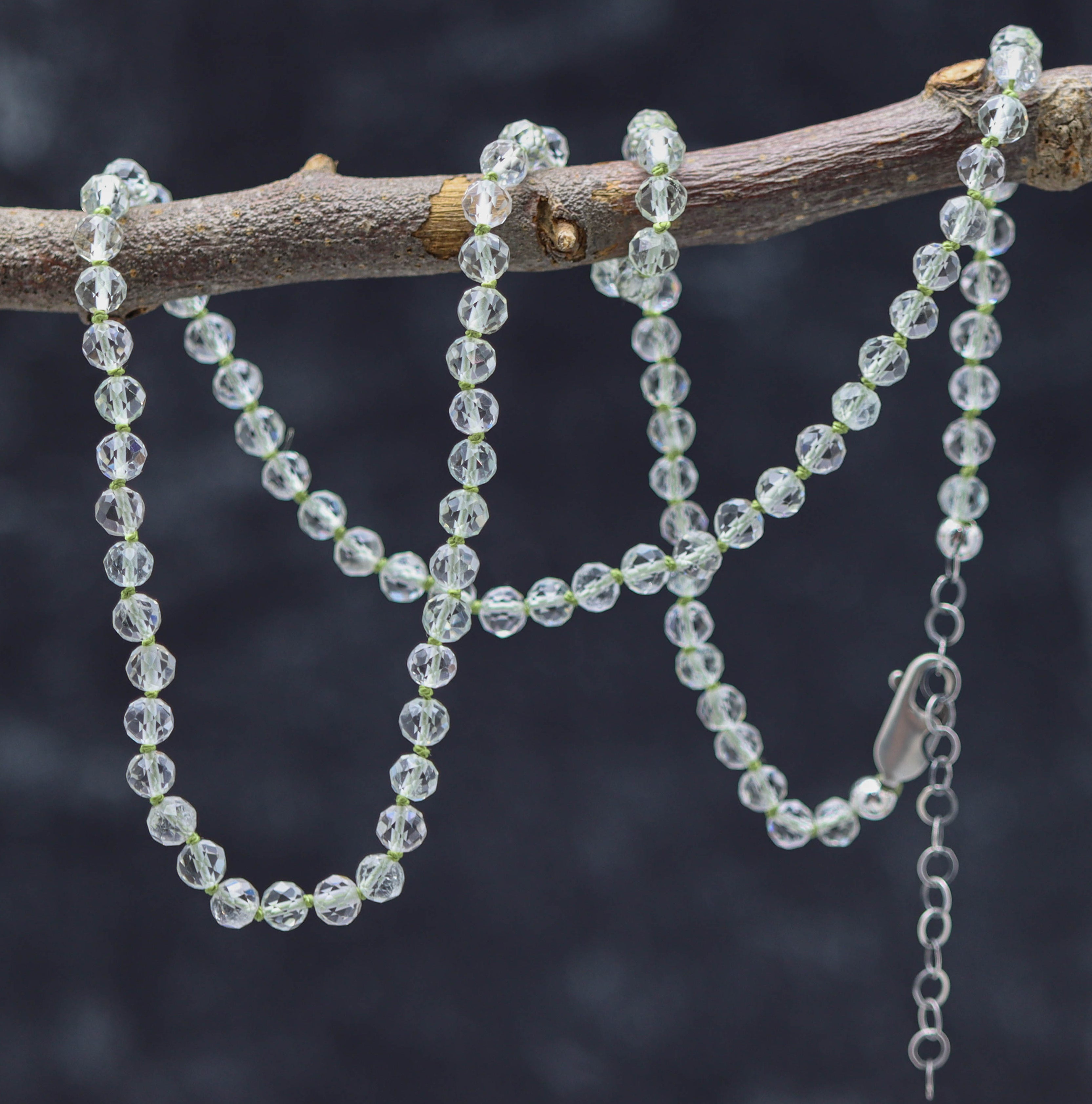 A sterling silver necklace with hand knotted green amethyst beads, displayed on a branch against a dark background.