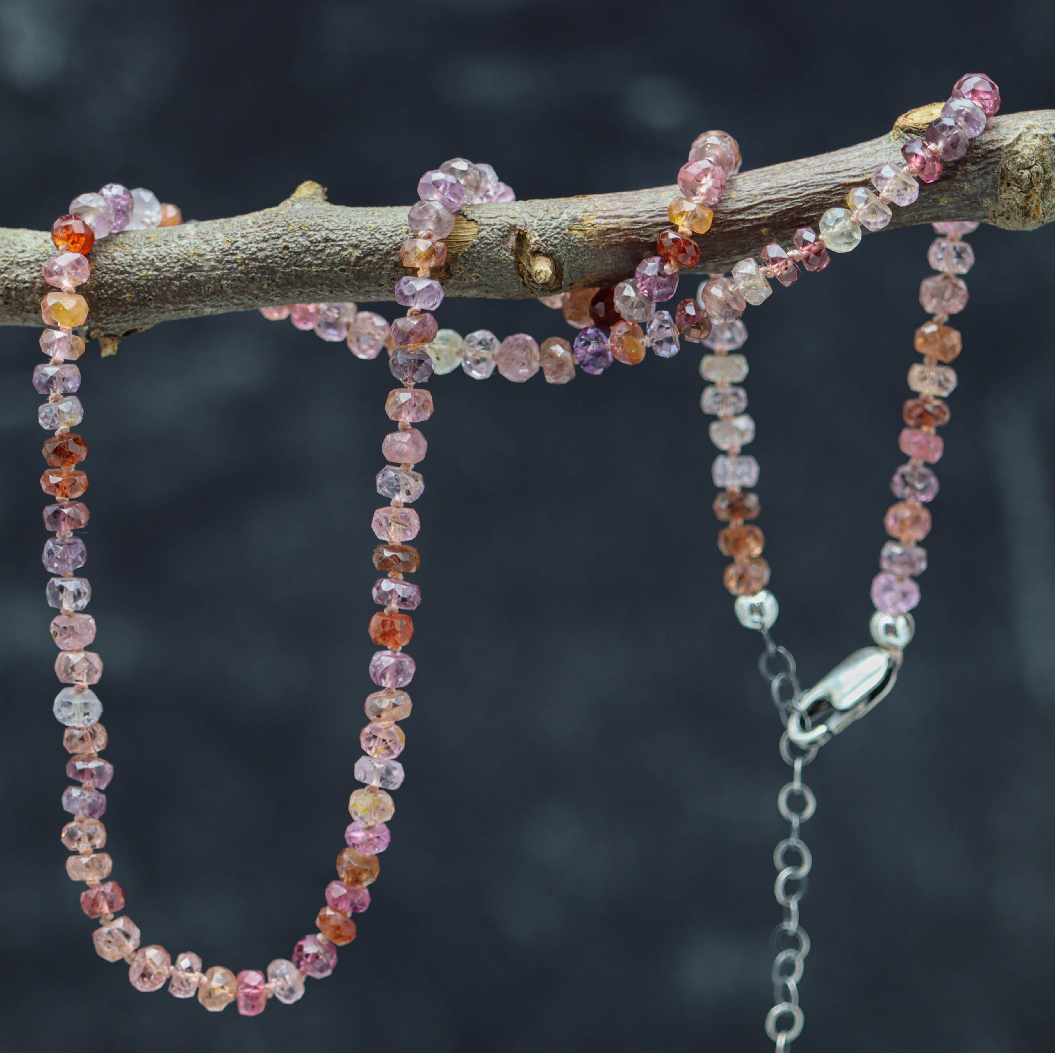 A hand knotted bead necklace made of multi-colored spinel stones on sterling silver, displayed on a branch.