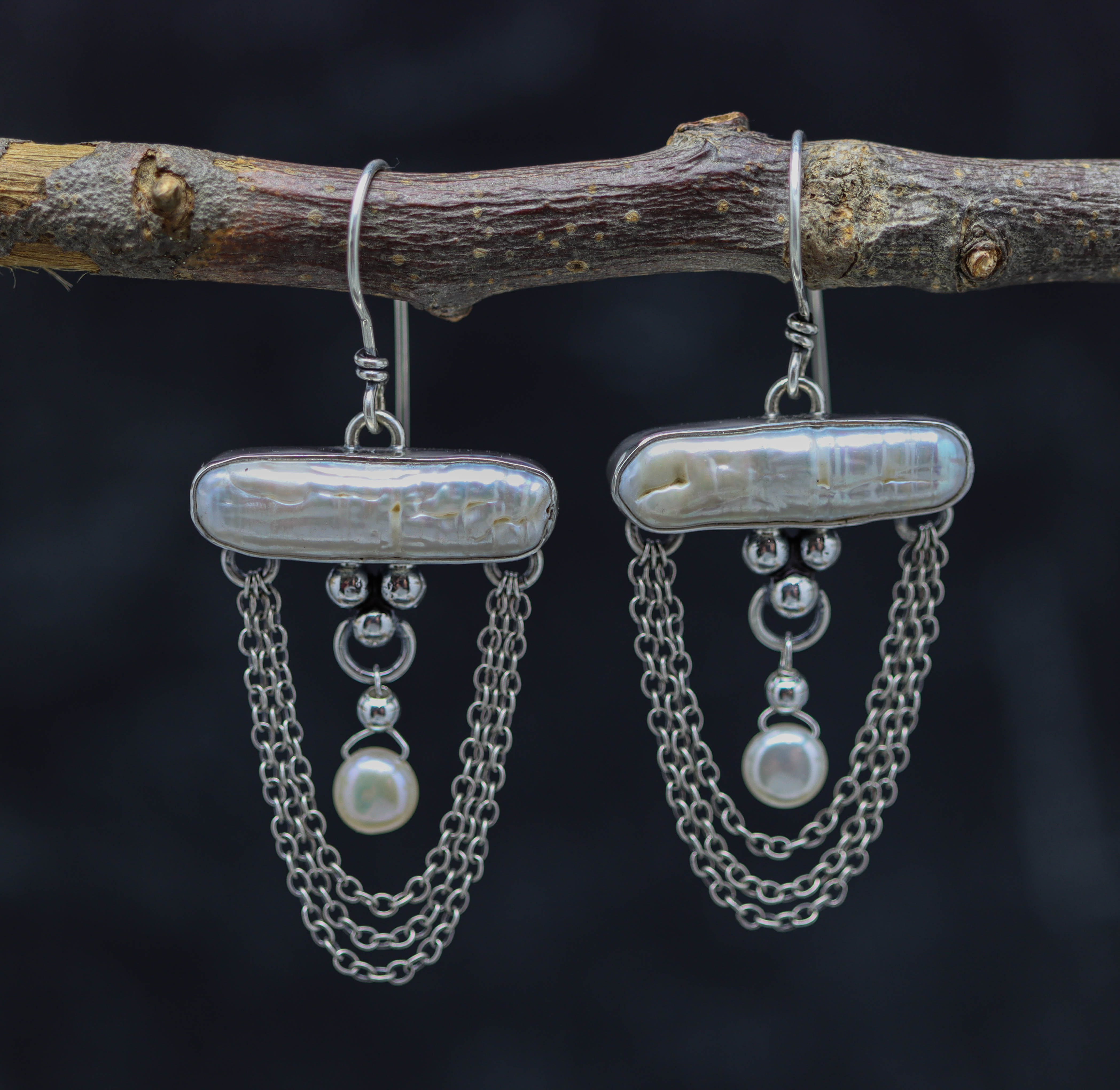 A pair of sterling silver earrings featuring oblong and flat round white pearls, with dangle silver chain components, displayed on a branch against a dark background.