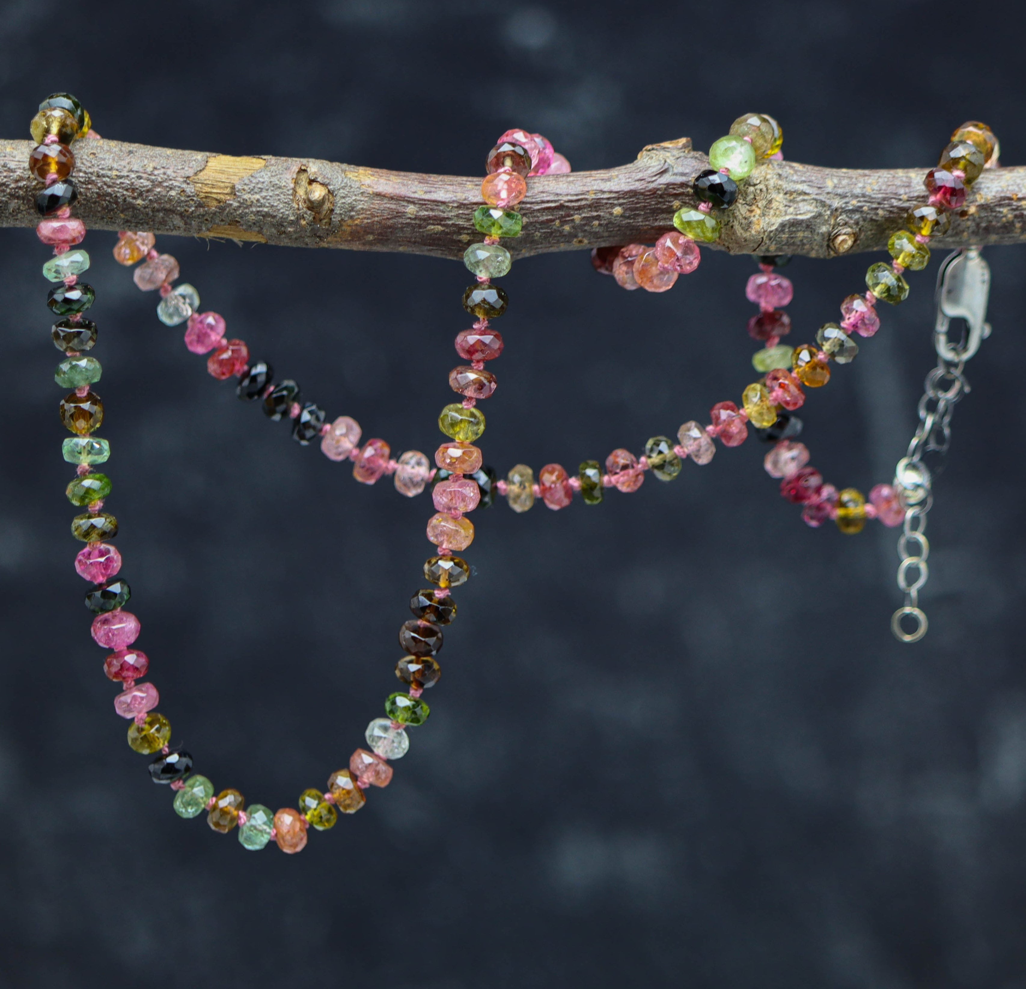 A hand knotted bead necklace made of multi-color tourmaline beads and sterling silver, displayed on a branch.