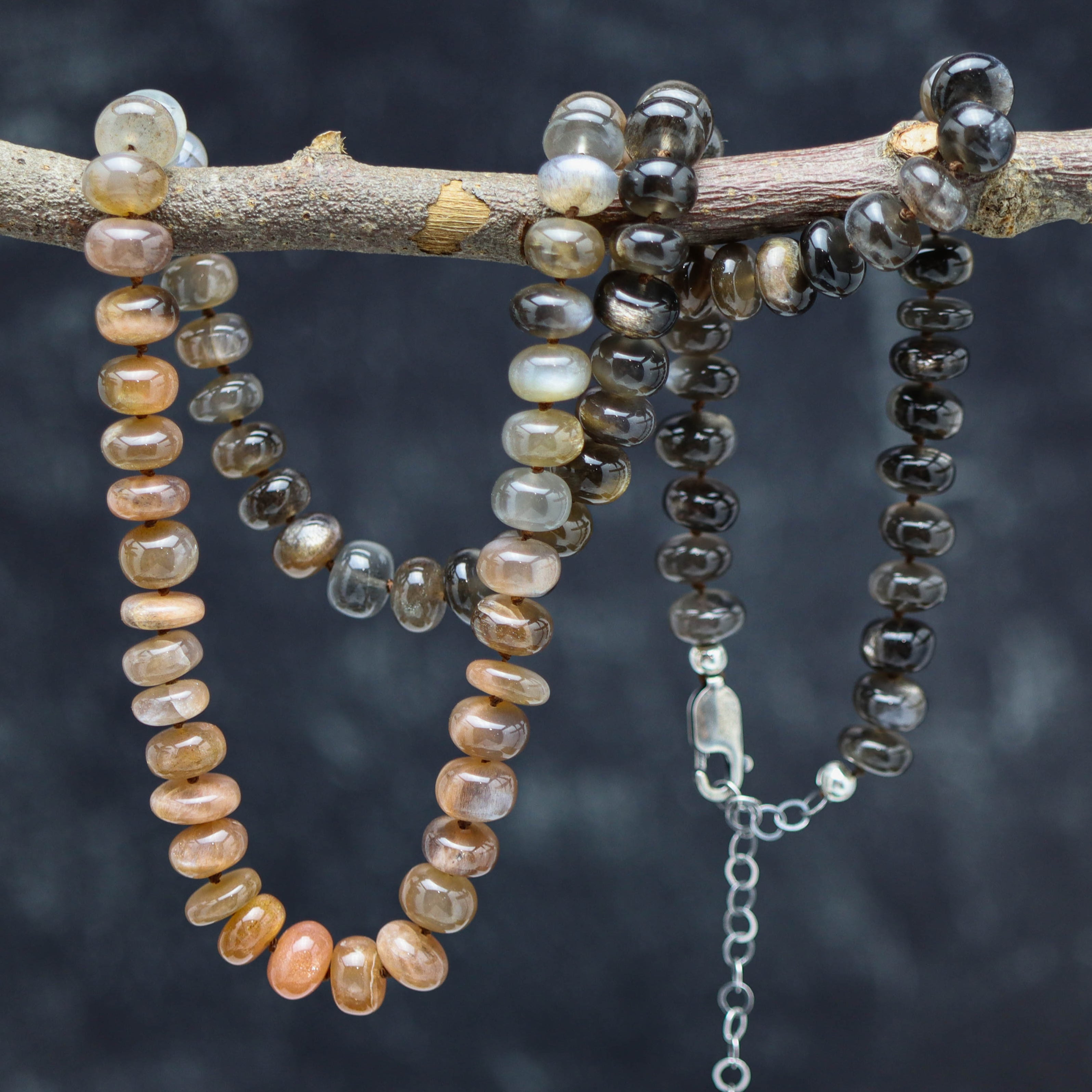 A hand knotted bead necklace with a sterling silver lobster claw closure, featuring a variety of moonstone beads in a gradient of colors from dark to light gray and tan.