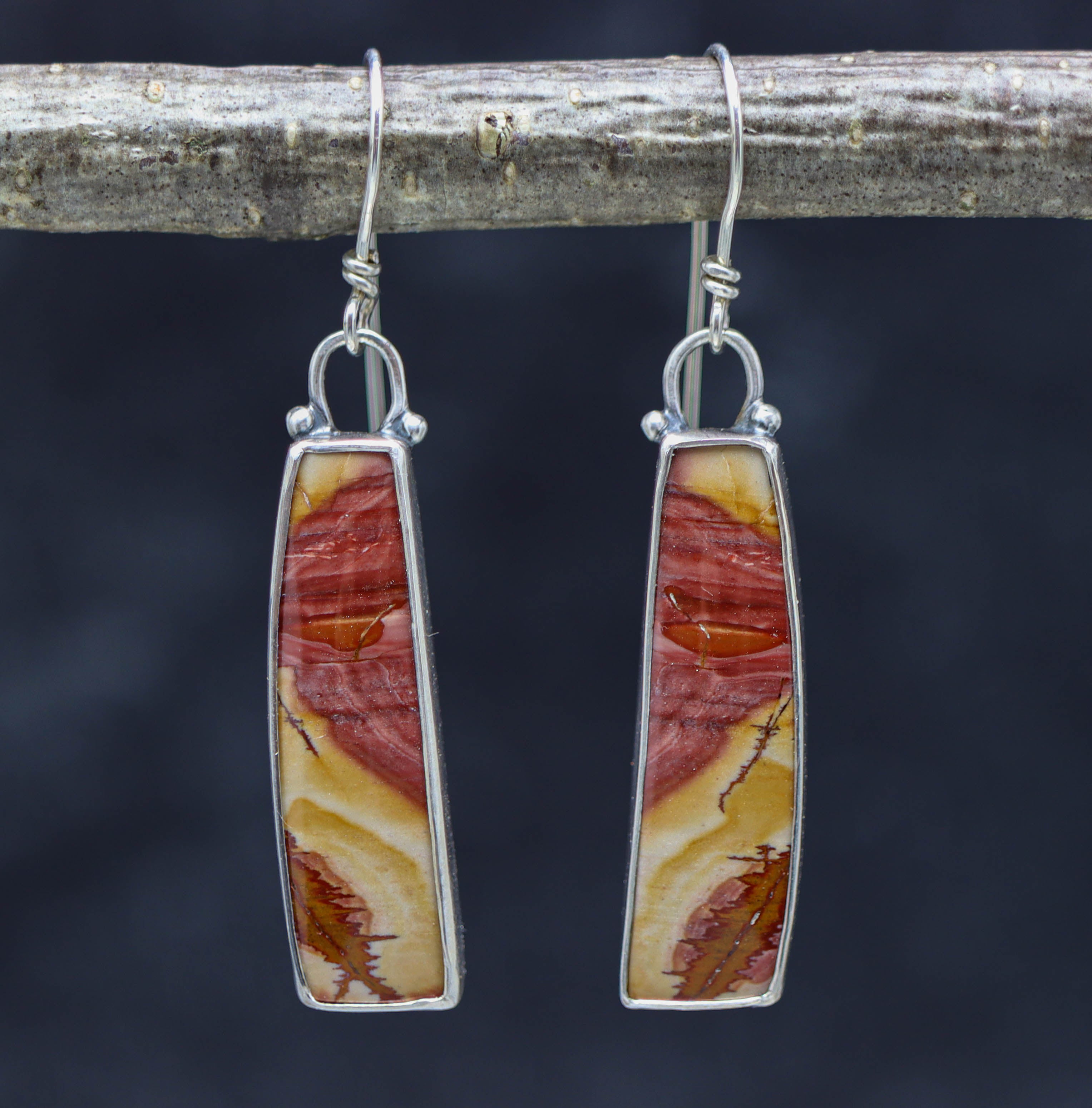 A pair of sterling silver dangle earrings with red falcon jasper setings, displayed against a dark background.