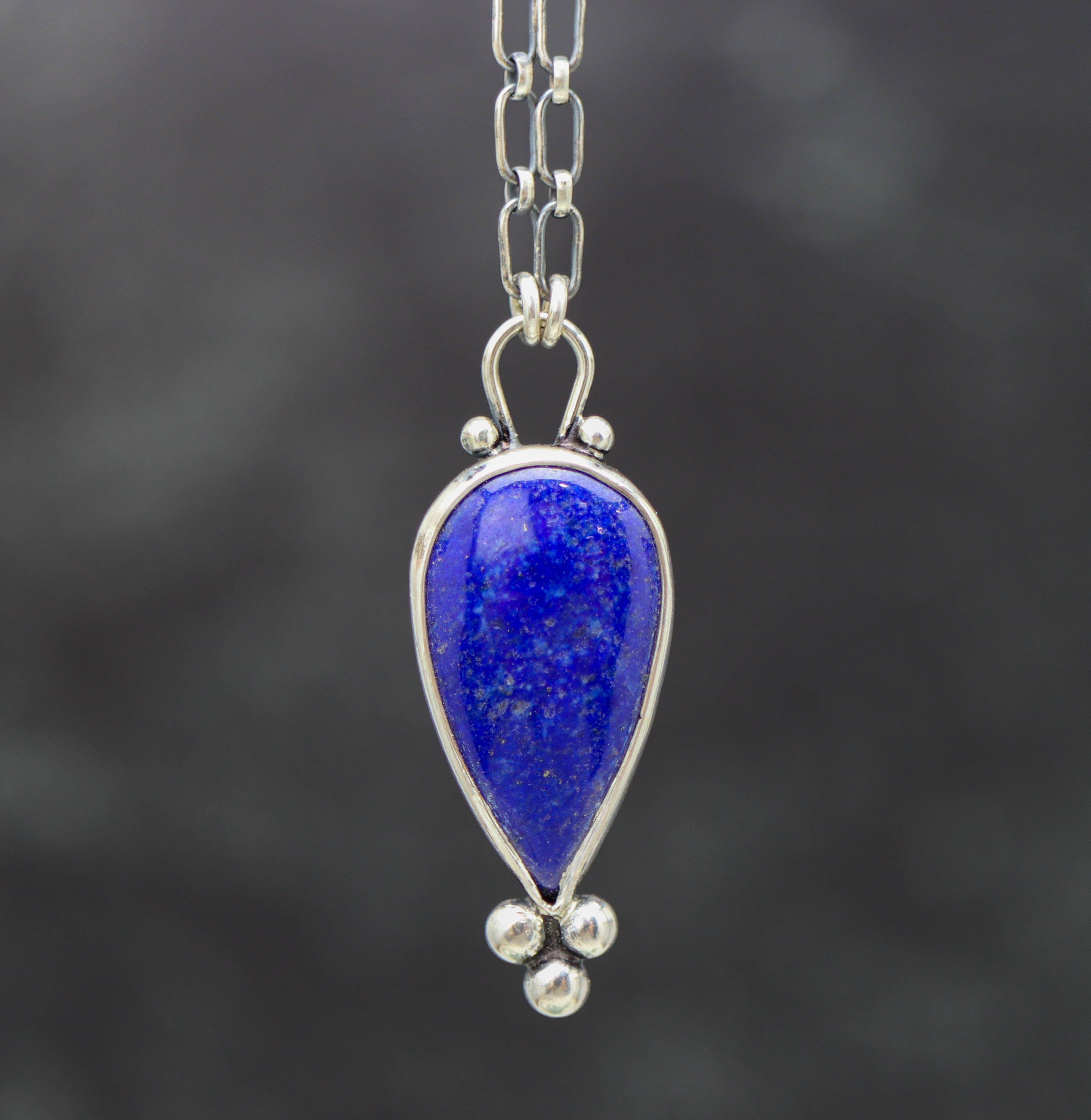 A sterling silver pendant necklace with a blue lapis lazuli gemstone centerpiece and small silver ball accents, featuring a lobster claw closure.