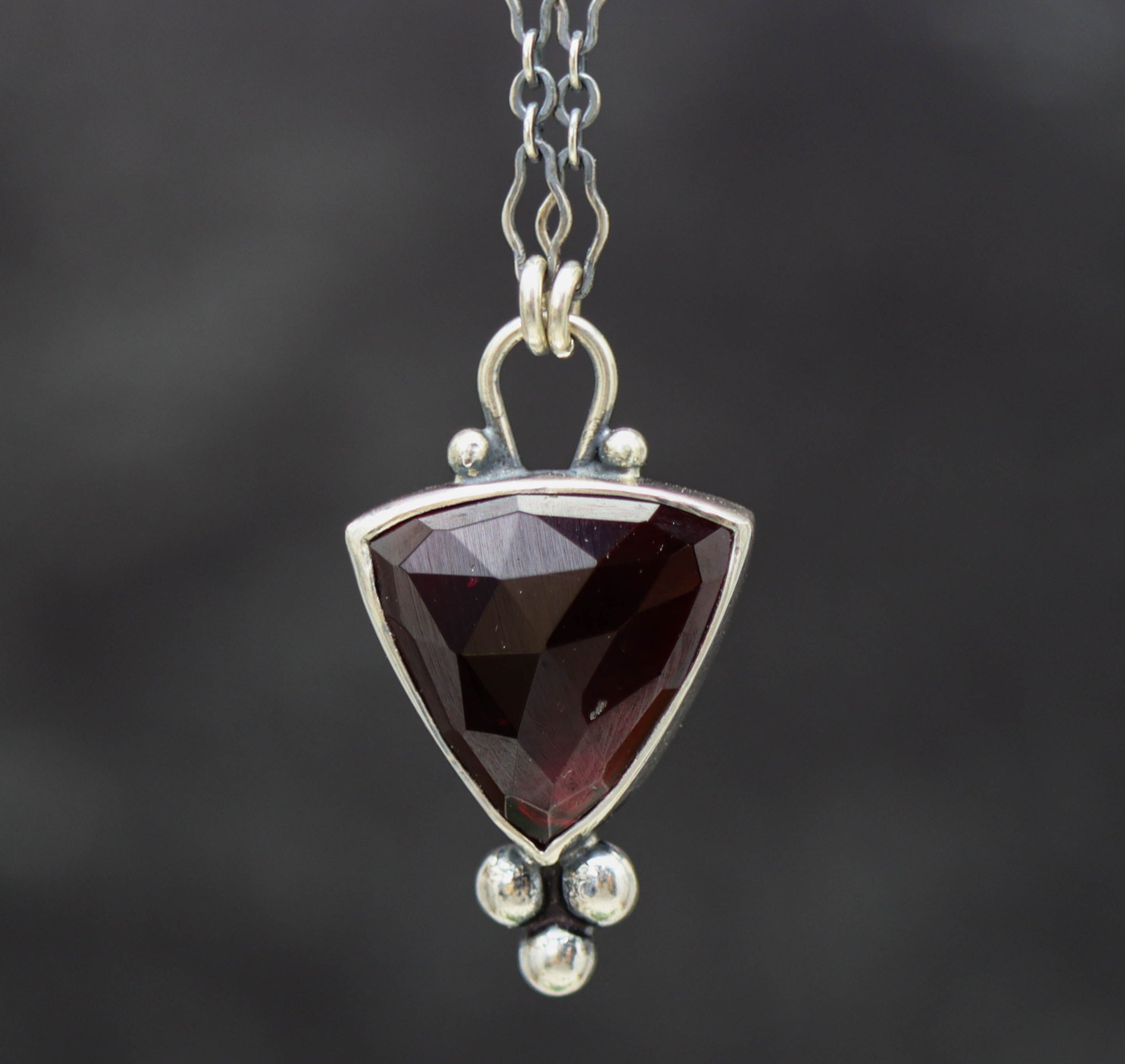 A sterling silver pendant necklace with a garnet centerpiece and small silver ball accents, featuring a textured backplate.