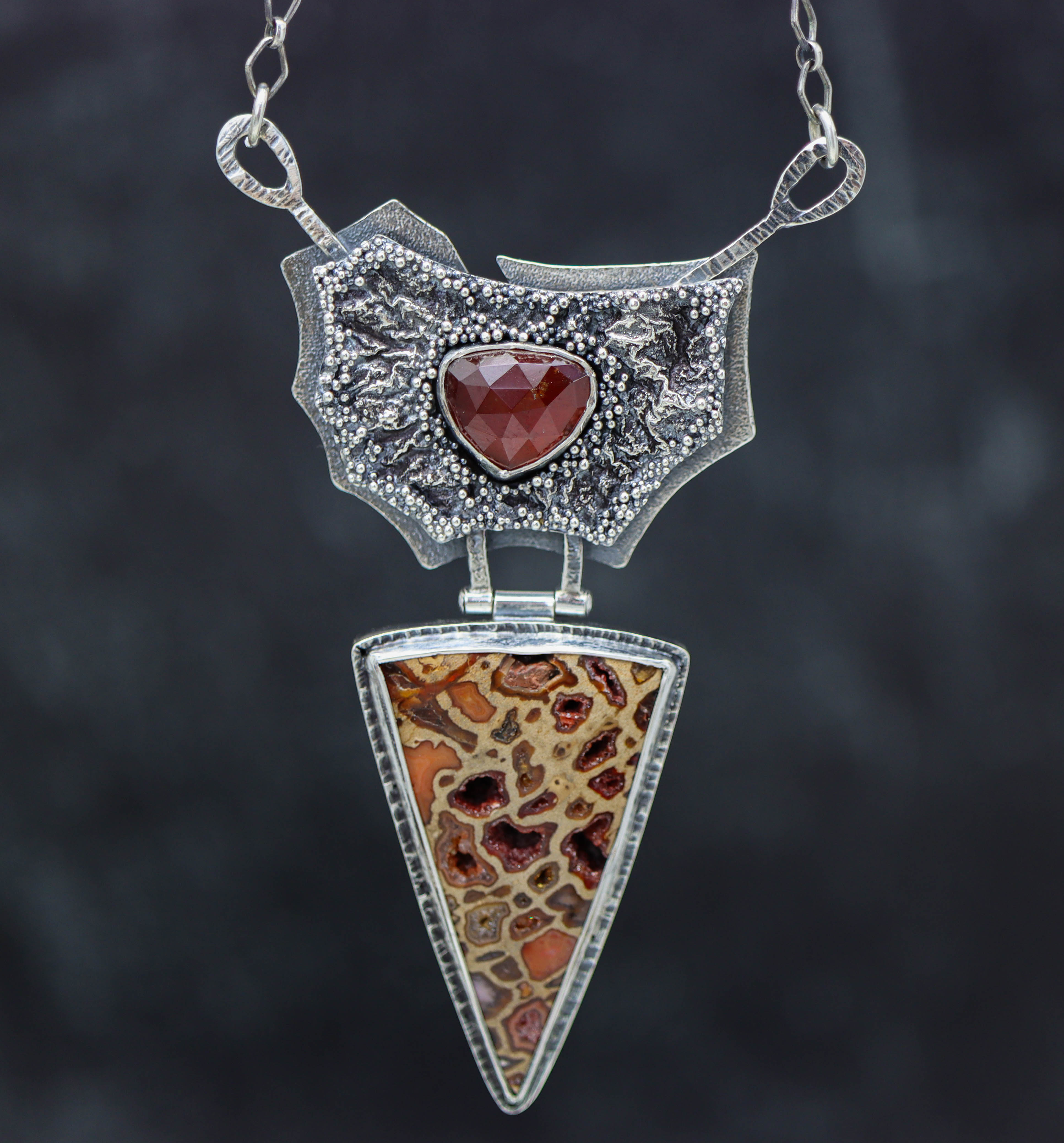 A sterling silver puzzle clasp necklace with a layered pendant featuring a red garnet centerpiece, textured silver, and granulated edges.