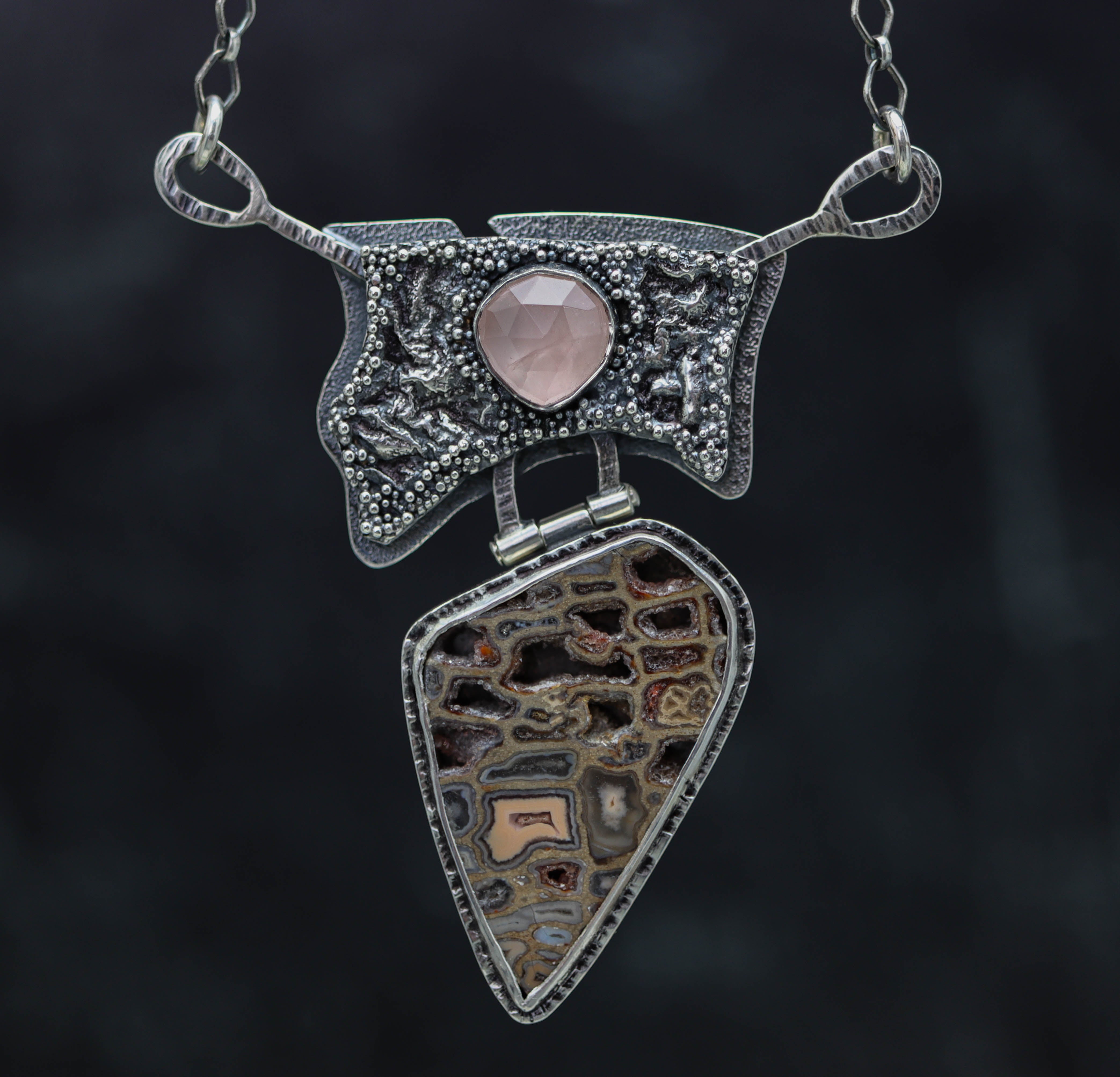 A sterling silver puzzle clasp necklace with a reticulation silver pendant featuring a rose quartz centerpiece and granulated edges.
