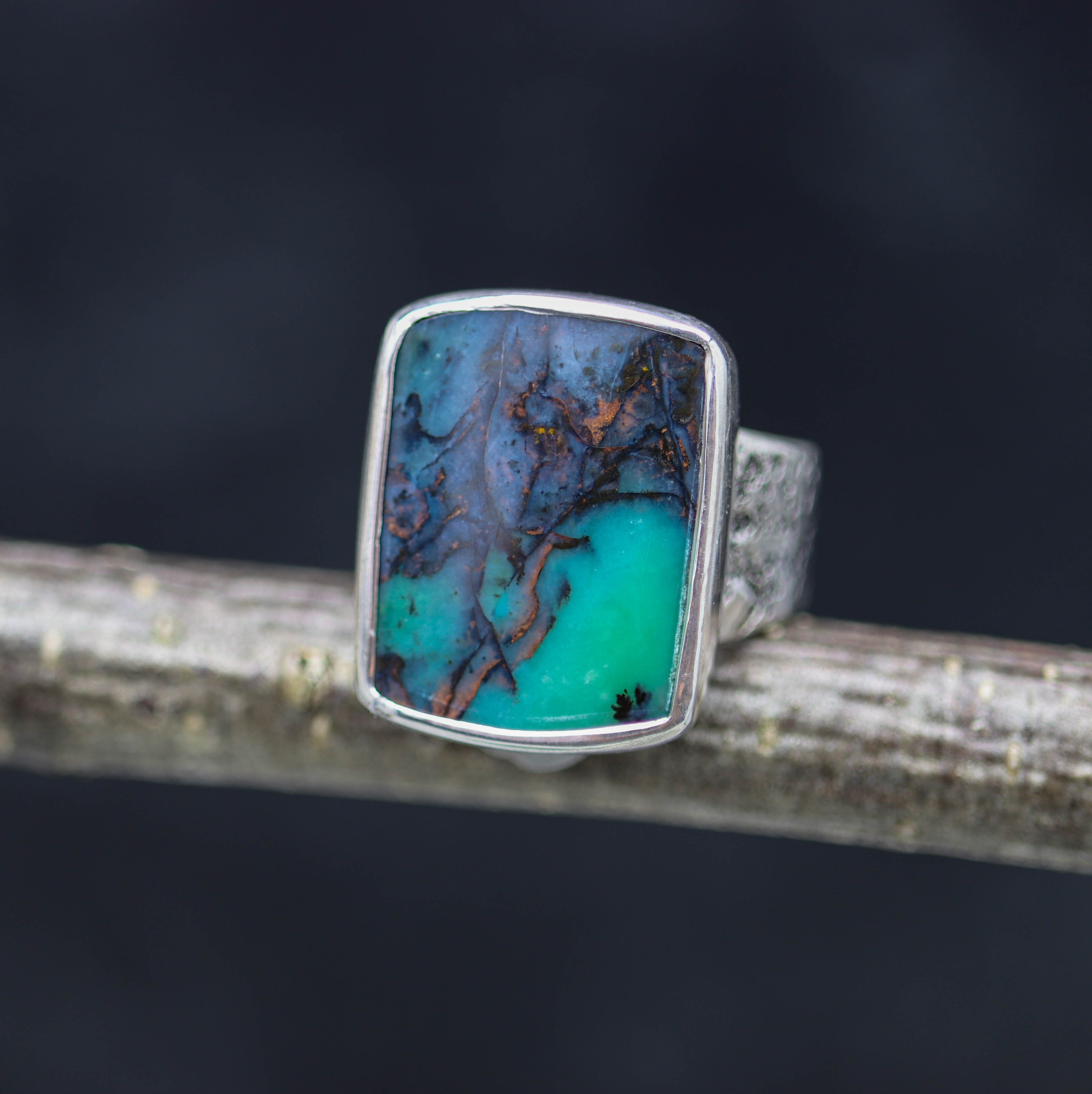 A sterling silver wide band ring with an opalized petrified wood stone set in the center, showcasing a blue green color with a webby black matrix and flecks of copper, placed on a wooden branch.