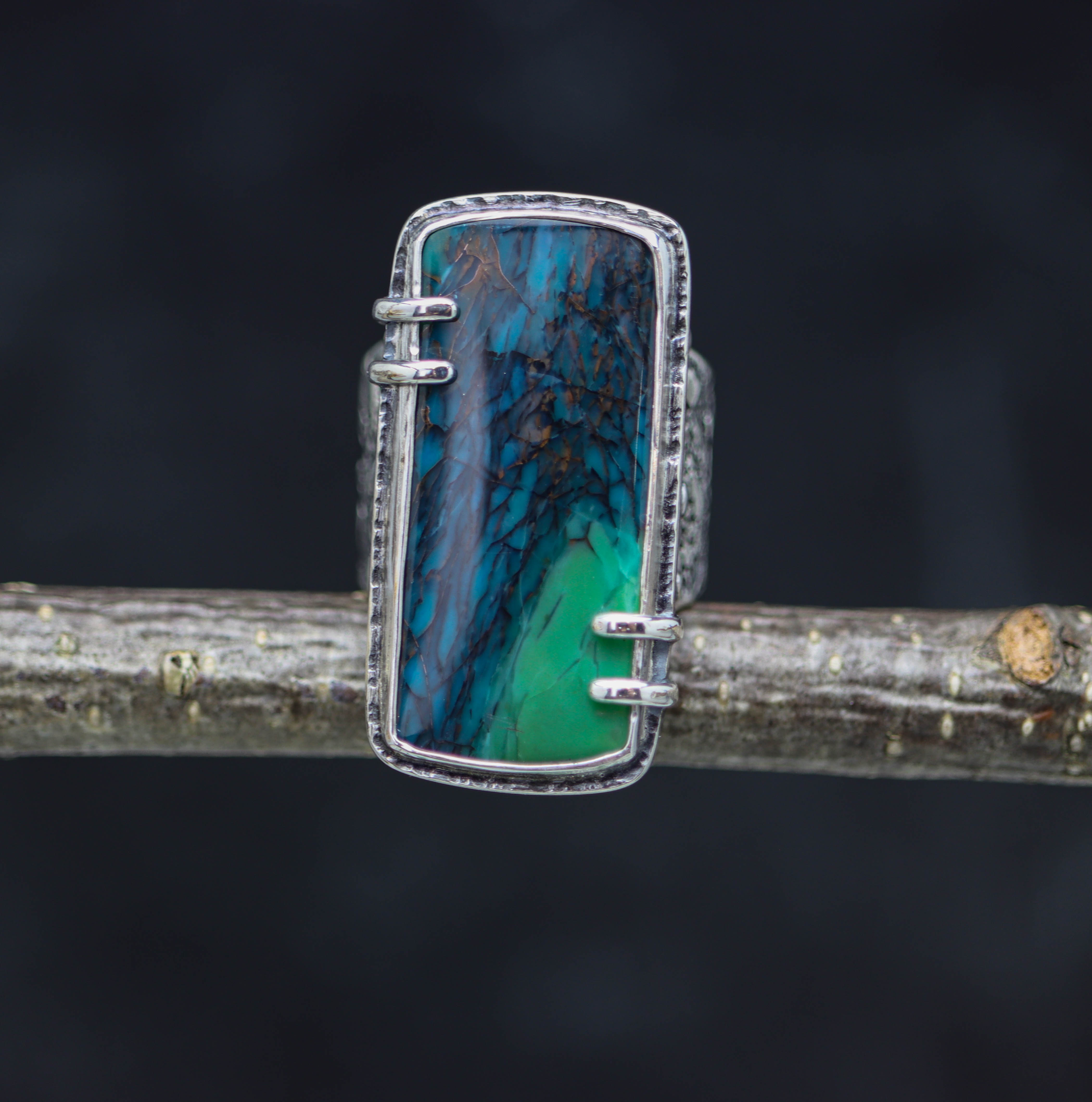 A wide band sterling silver ring with a petrified opal wood stone set in a double bezel, showcasing blue green color with black matrix and copper flecks.