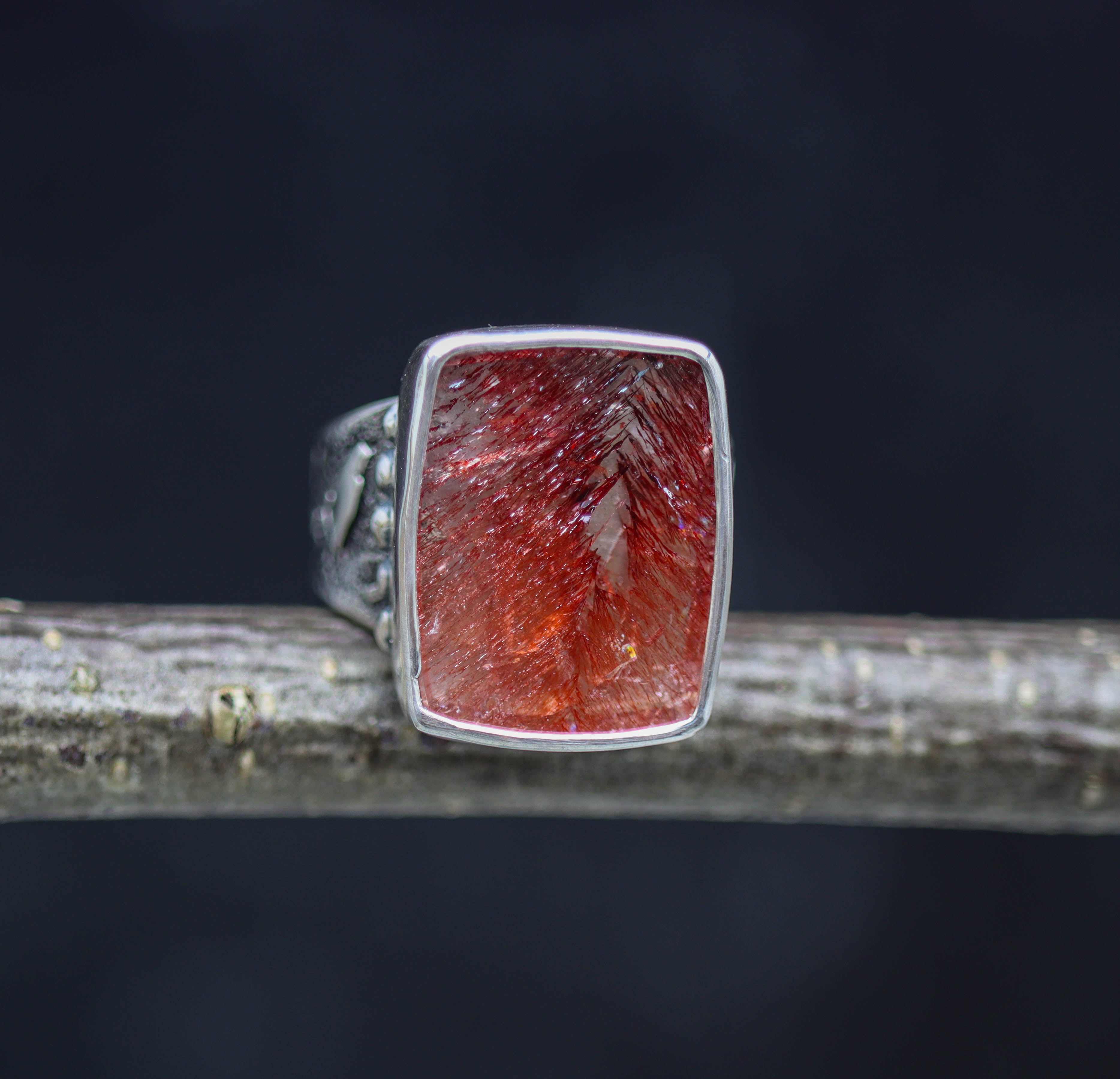 A sterling silver wide band ring with a rectangular red super seven gemstone set in a textured mount, displayed on a wooden branch against a black background.