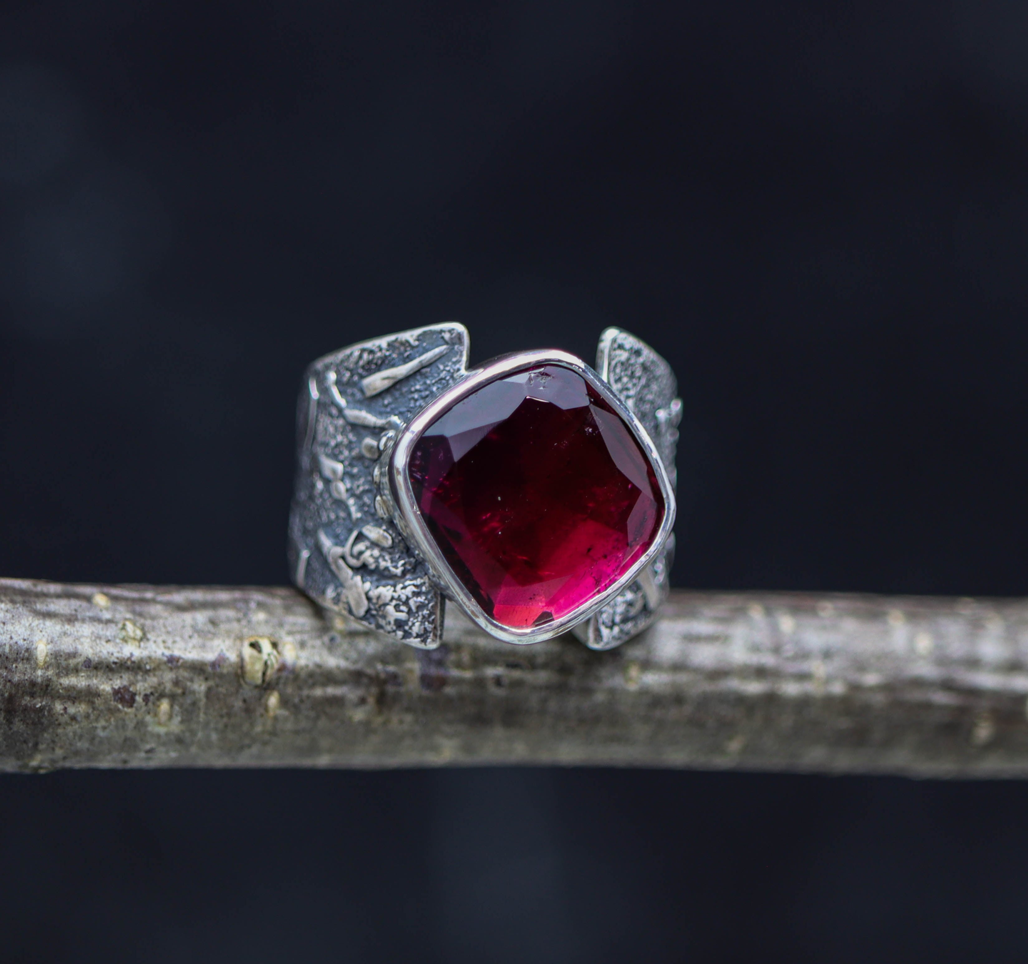 A sterling silver wide band ring with a large rubelite pink tourmaline rose cut, displayed on a wooden branch against a dark background.
