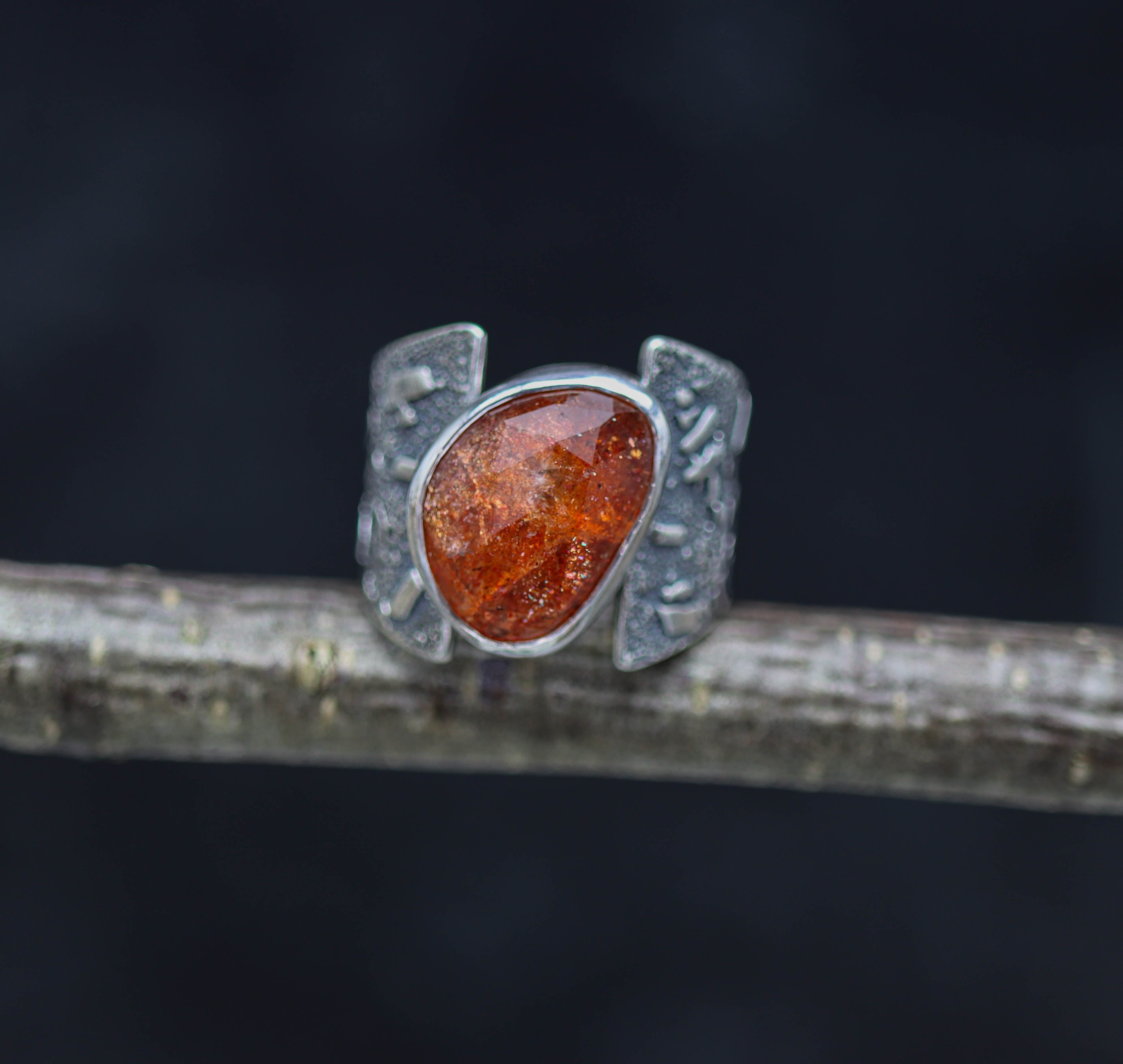 A sterling silver wide band ring with a rose cut sunstone set on a dark background, appearing orange with a textured design.