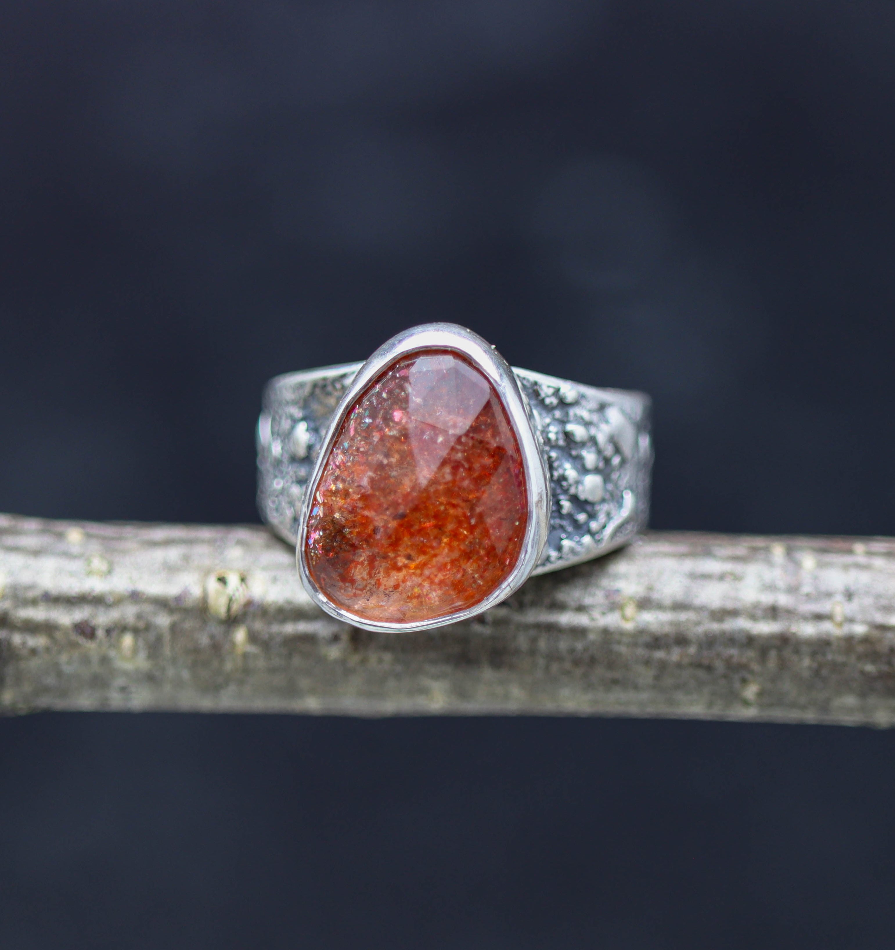 A sterling silver wide band ring with a rose cut sunstone set on a dark, textured surface.