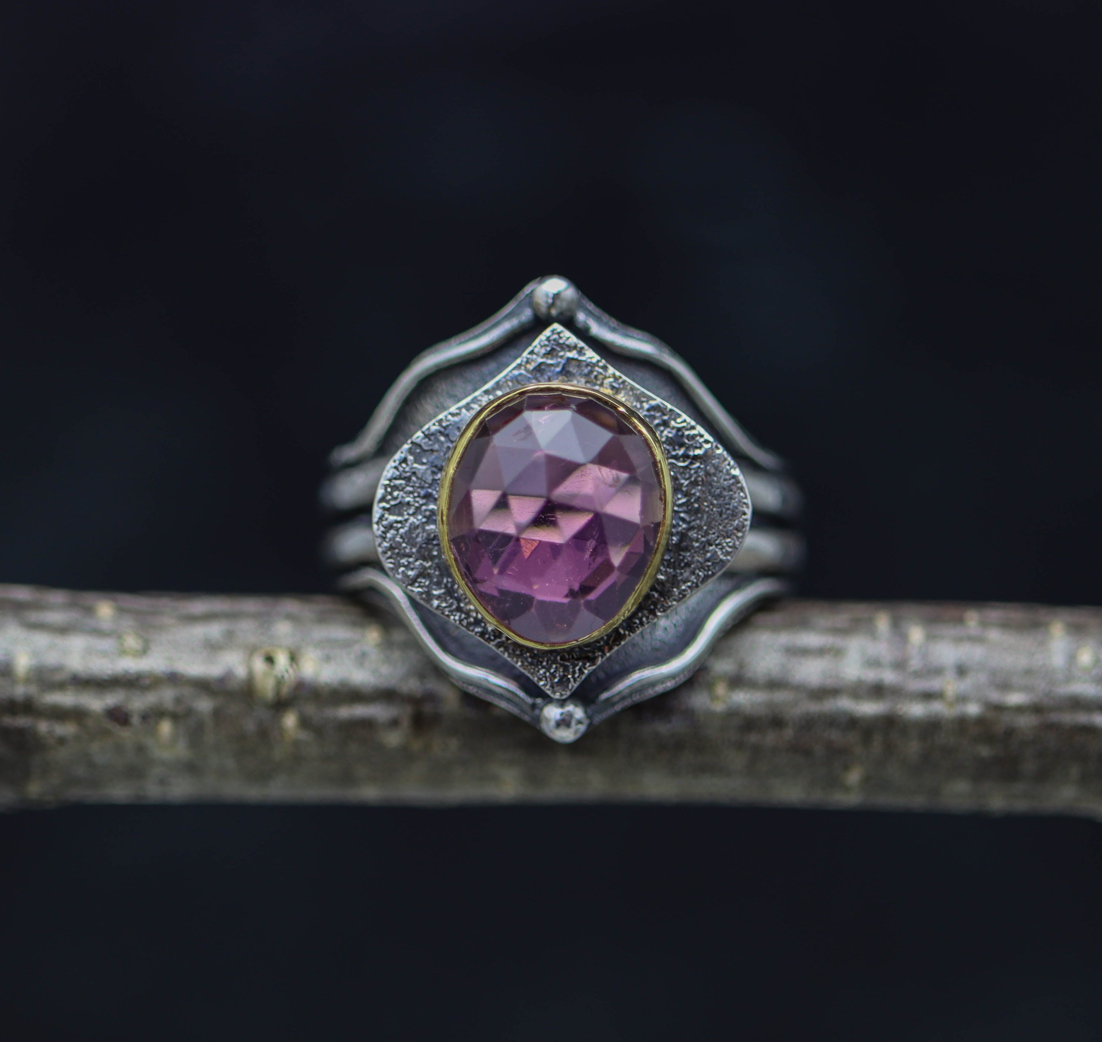 A platform ring with a pink tourmaline stone, set in a sterling silver and 22k gold setting, displayed on a wooden branch against a black background.