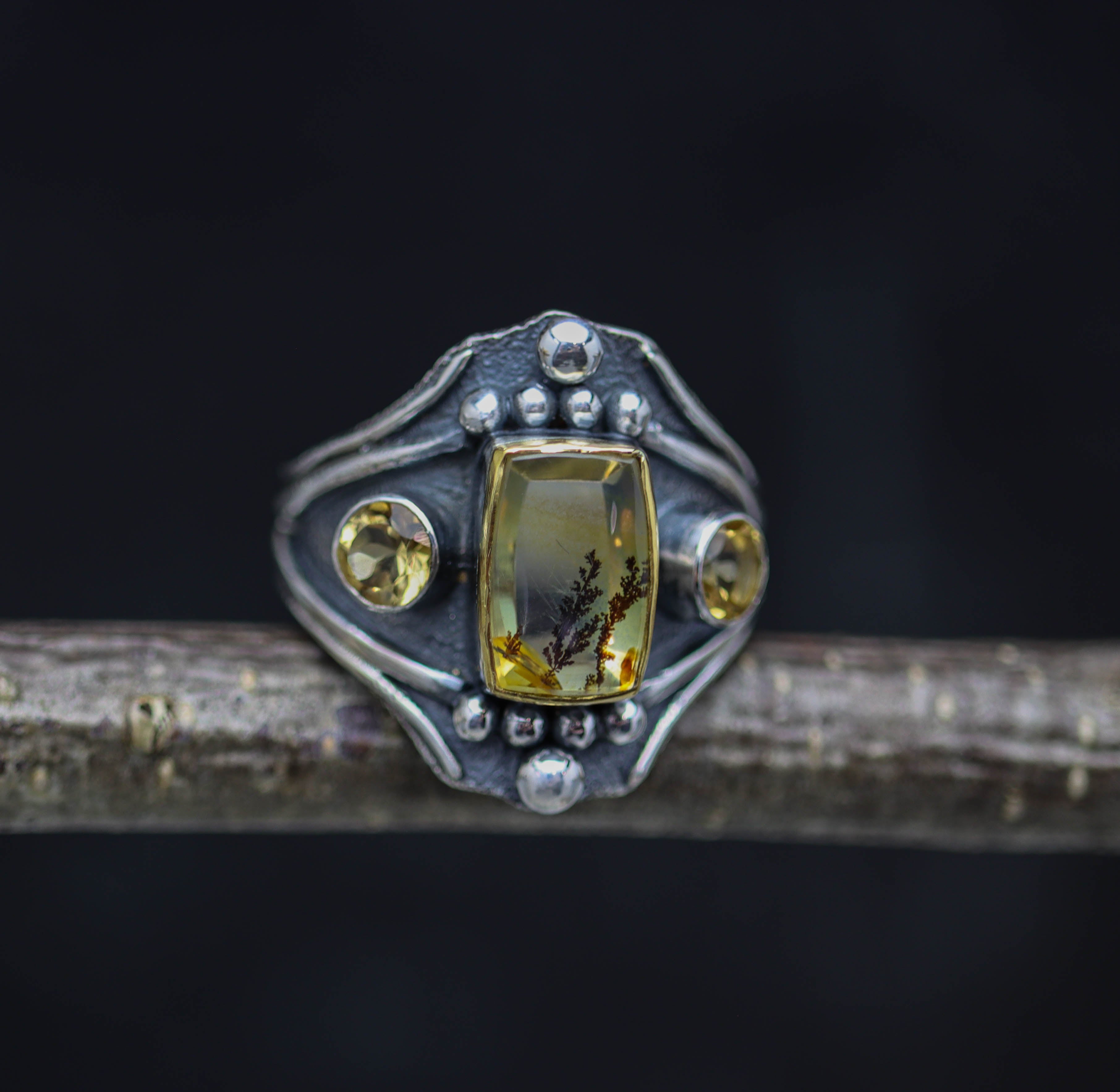 A sterling silver and 22k gold ring with a dendritic quartz center stone and two faceted yellow citrines on a dark background.