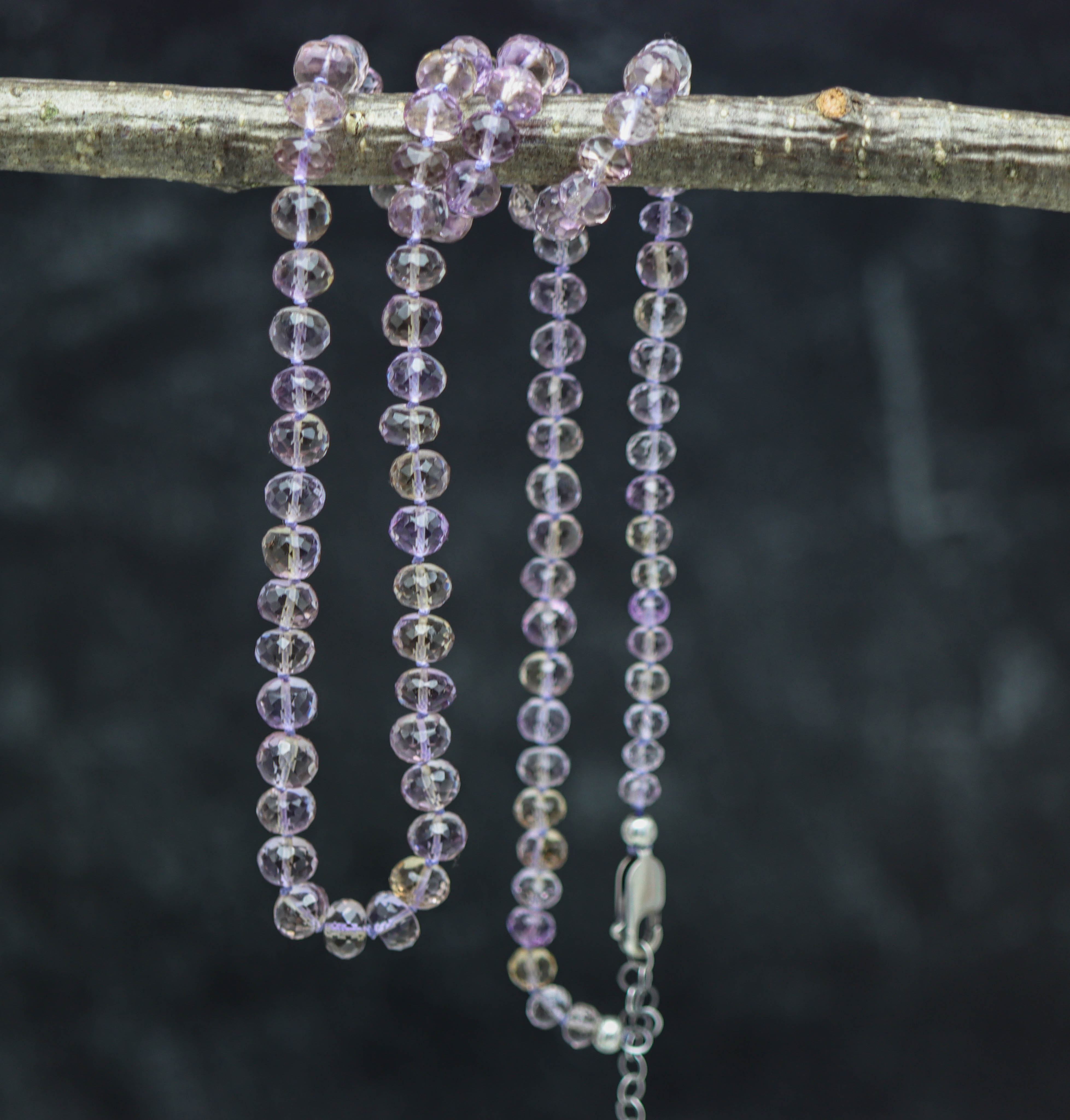 A hand knotted beaded necklace made of ametrine stones, with a sterling silver clasp, displayed on a branch against a dark background.