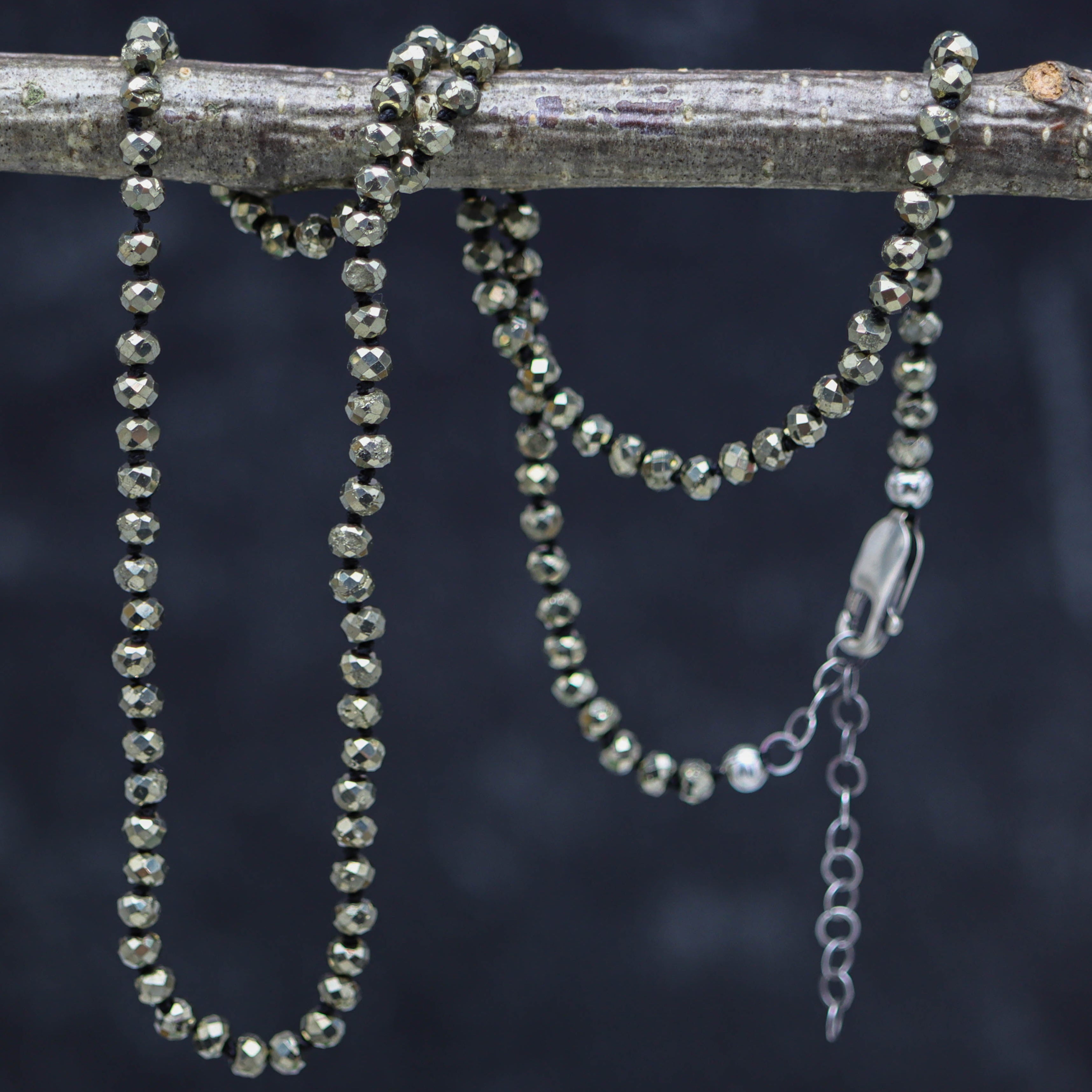A hand knotted bead necklace made of faceted pyrite roundels and sterling silver, displayed on a branch against a dark background.