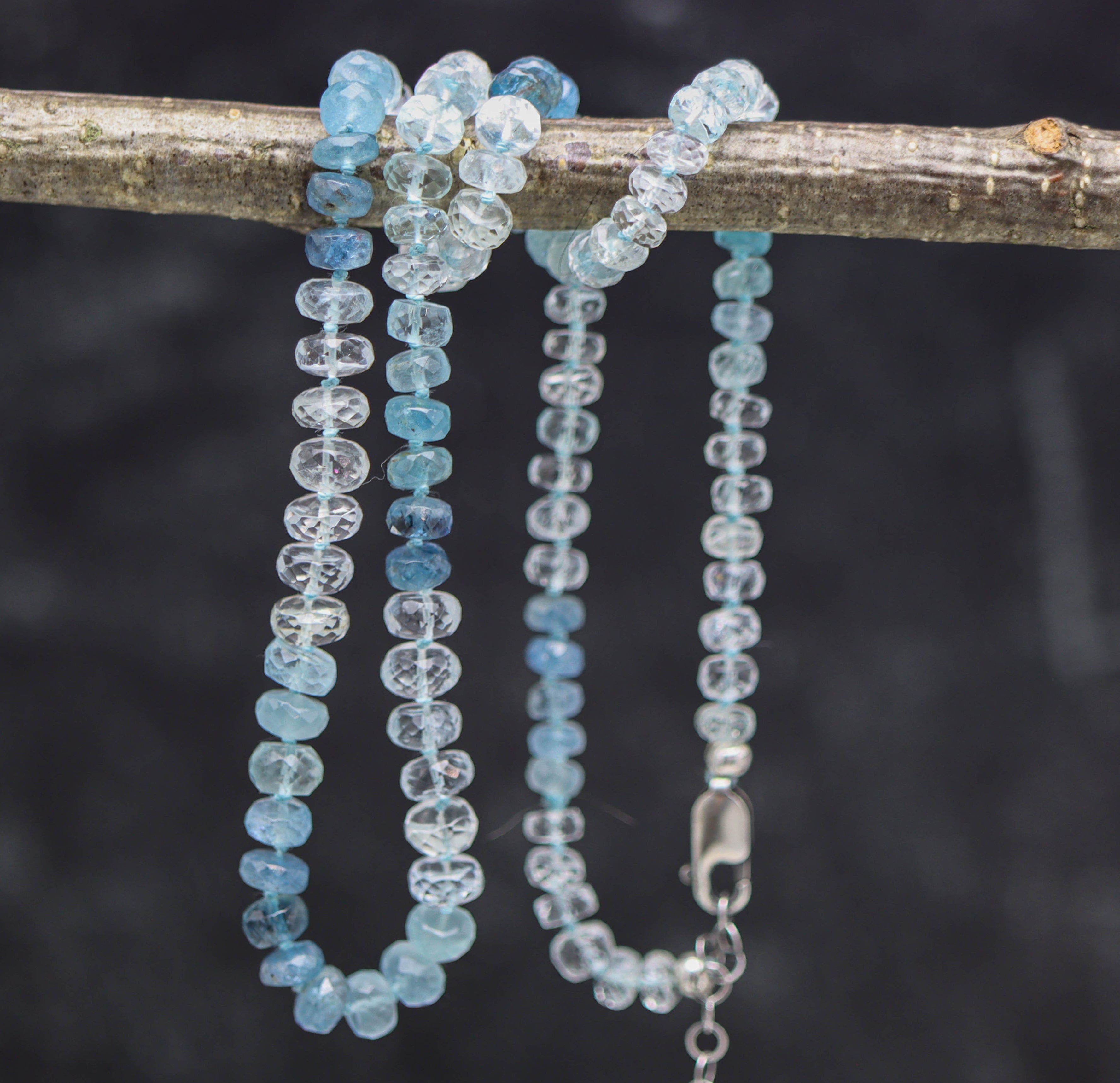A hand knotted bead necklace made of blue aquamarine roundels, with a sterling silver clasp, displayed on a branch against a dark background.