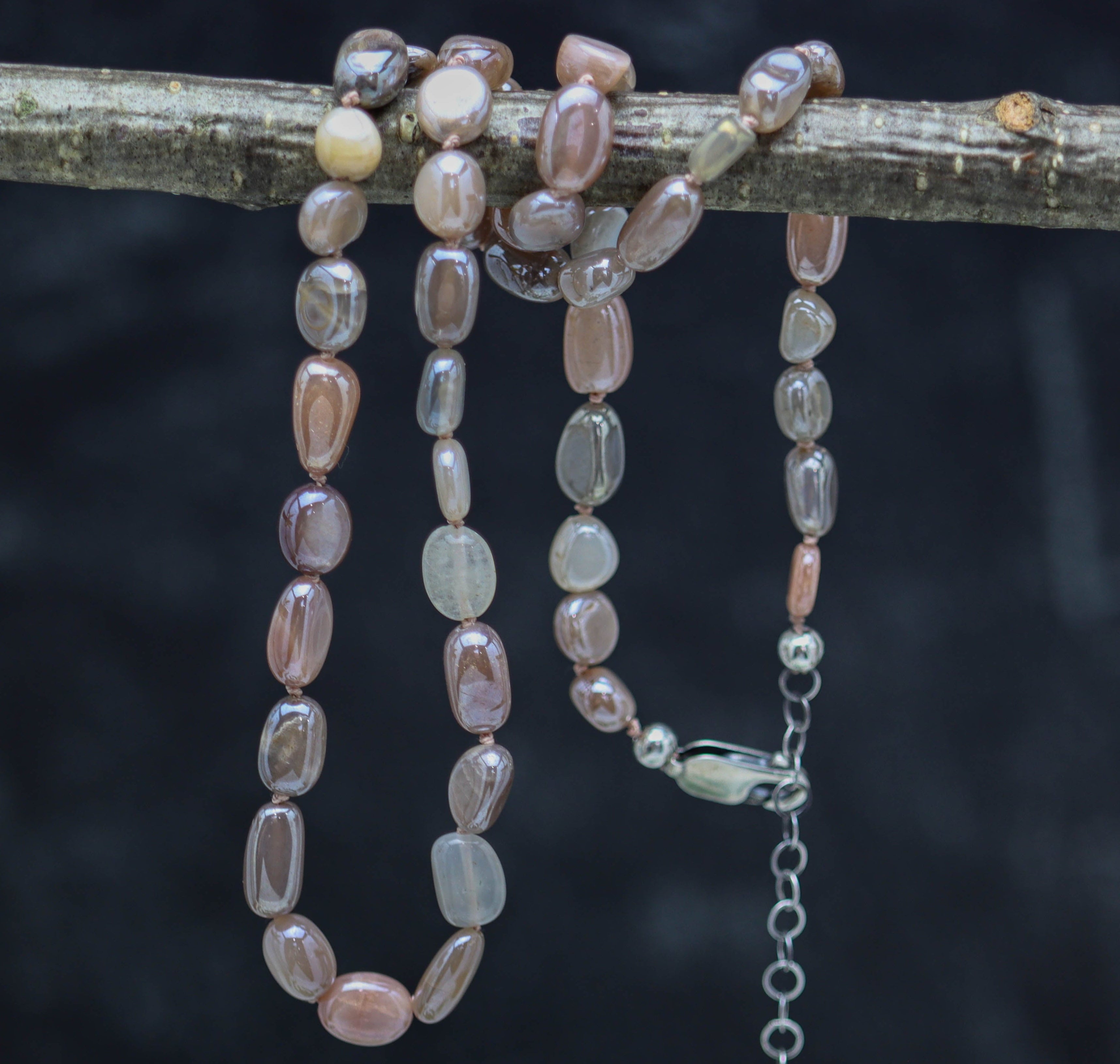A hand knotted bead necklace with sterling silver closure, featuring silvery flash moonstone beads in pinkish peach to grey to almost white shades, displayed on a dark background.