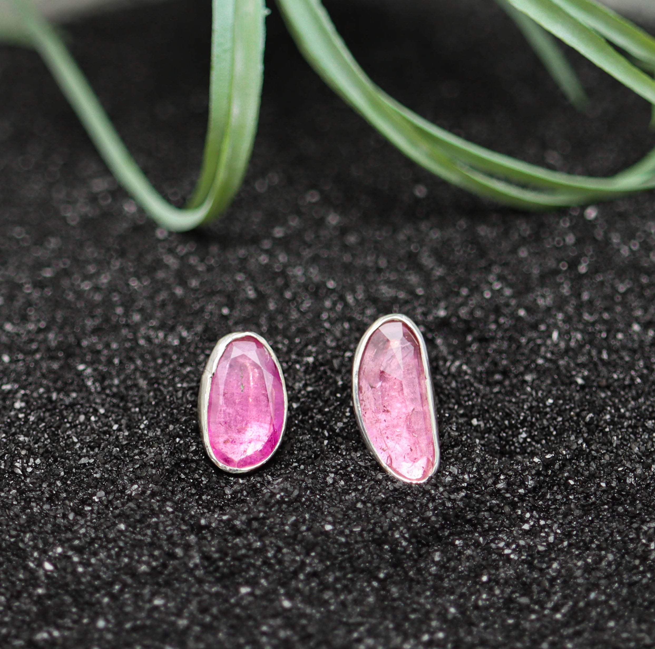 Pink gemstone earrings on a black glittery surface with green plant leaves.