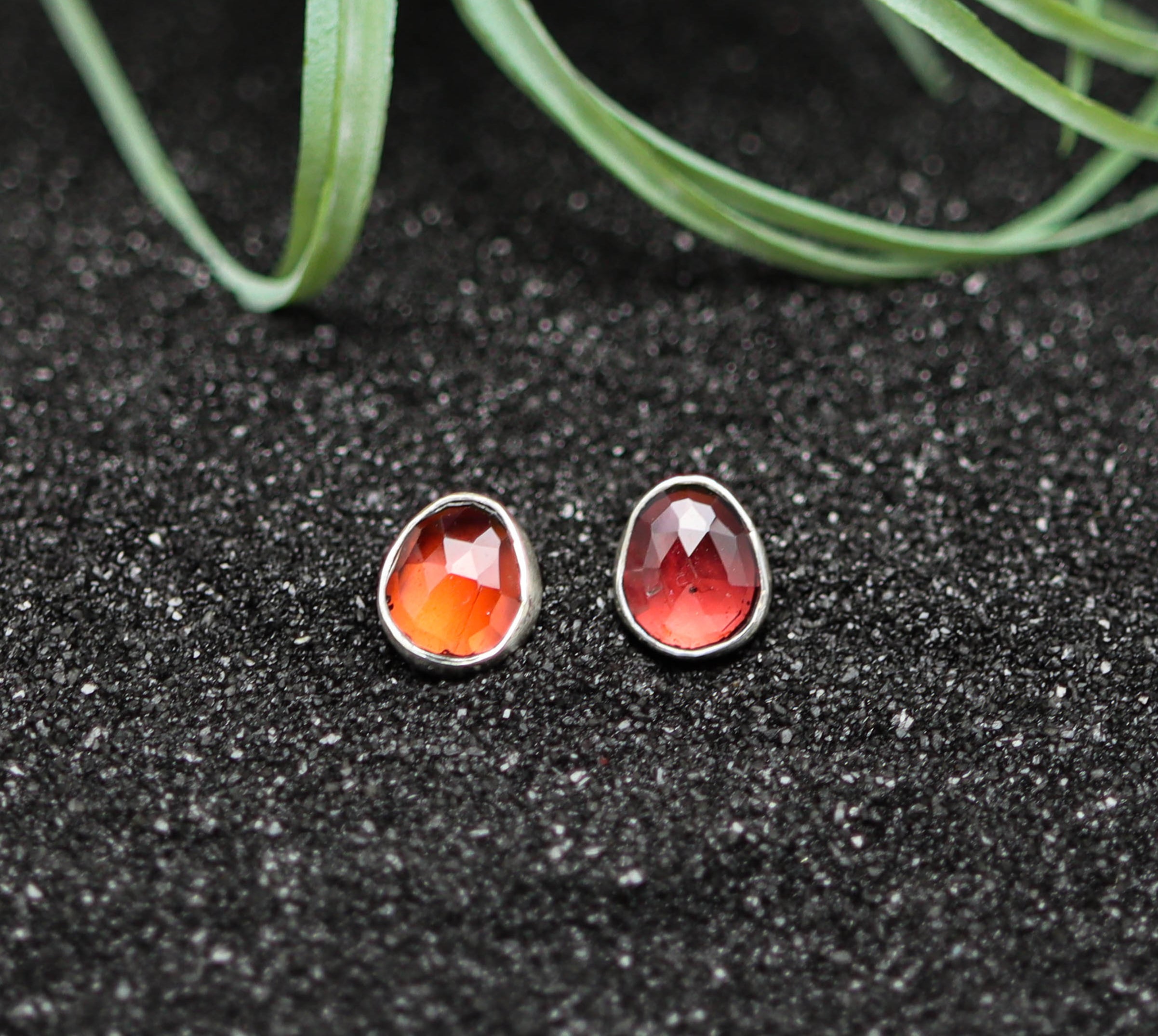 Teardrop-shaped red gemstone earrings on a black sand-like surface with a plant in the background.
