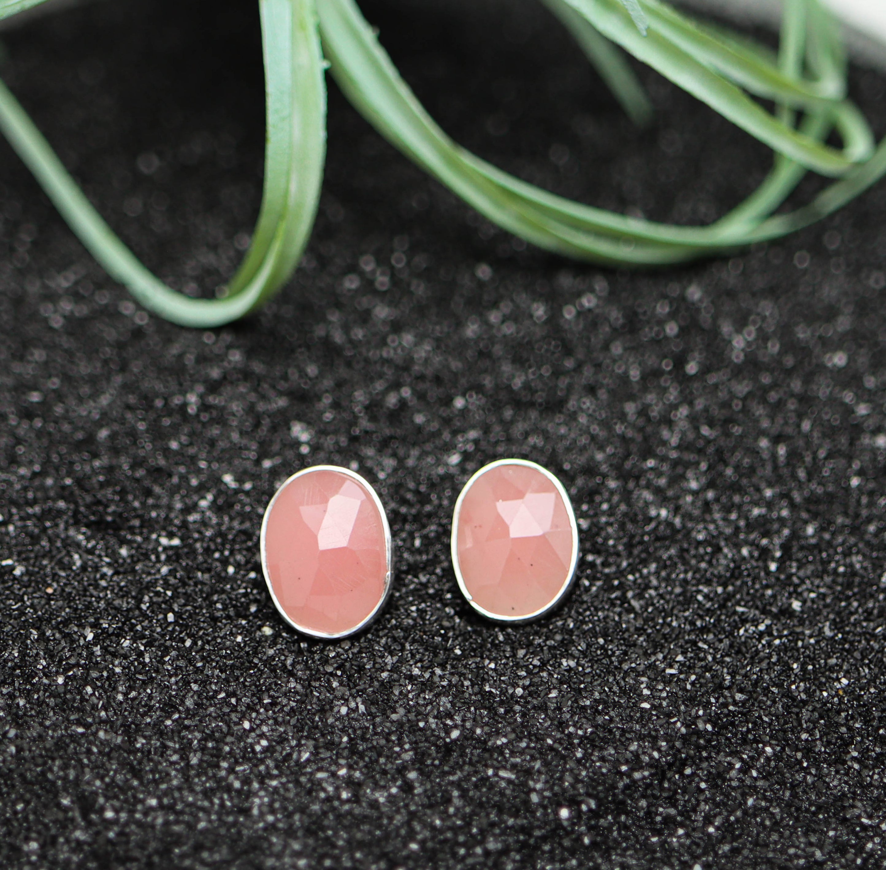 Pair of pink oval earrings on a black textured surface with green leaves in the background.
