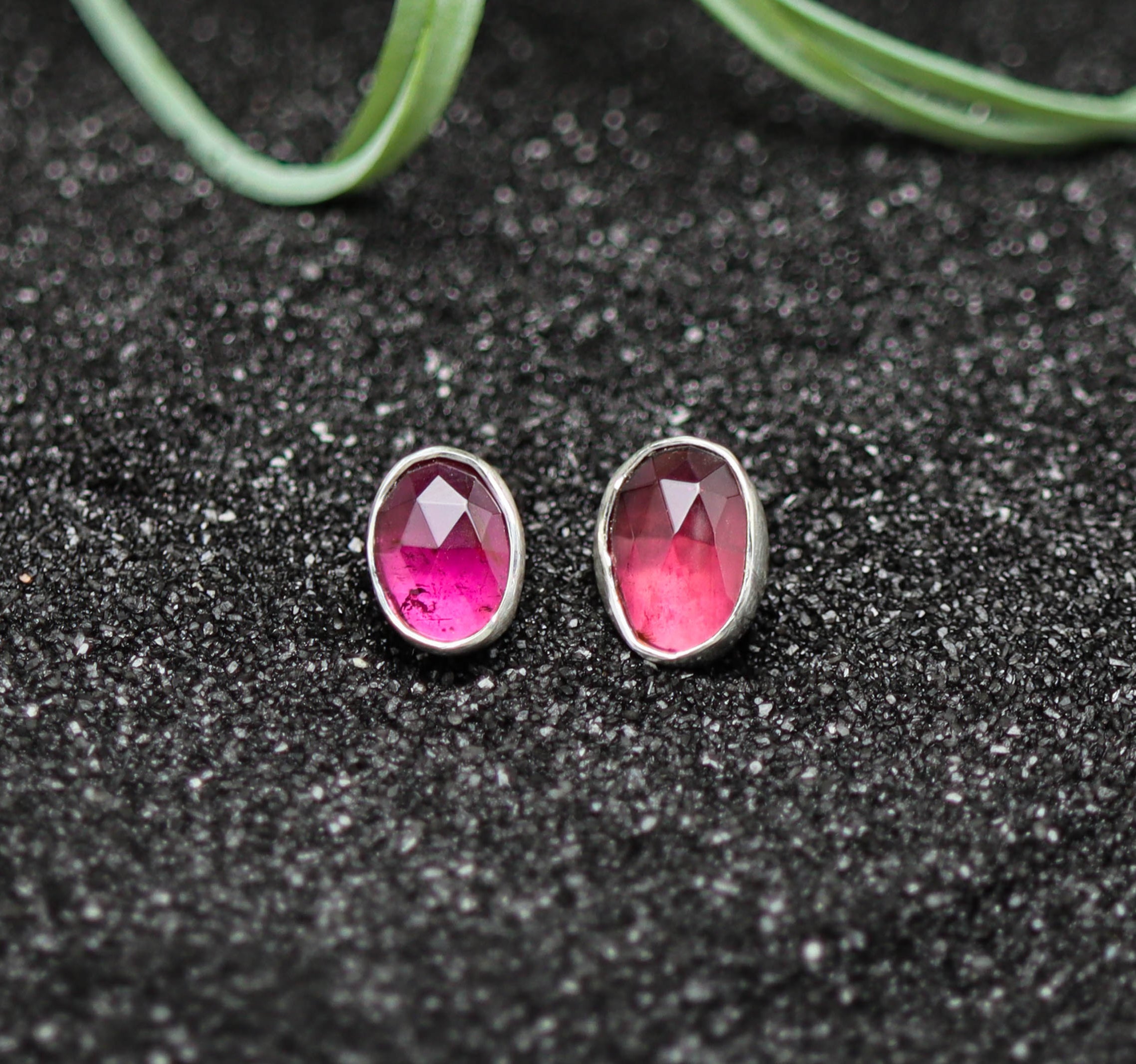 Pair of pink gemstone stud earrings on a black textured surface with green leaves in the background.