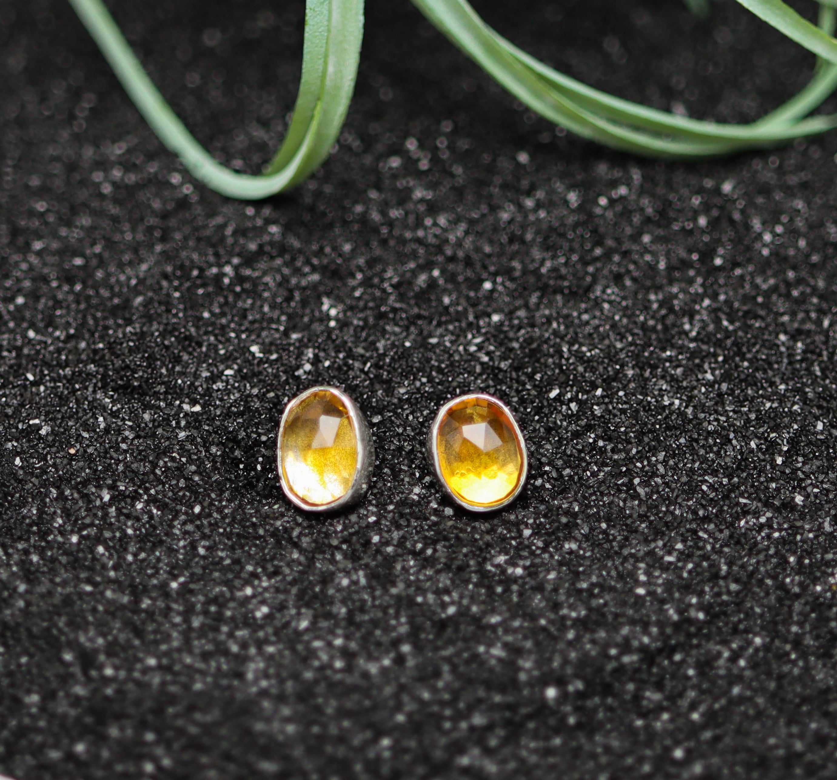 Pair of yellow citrine stud earrings on a black glittery surface with greenery.