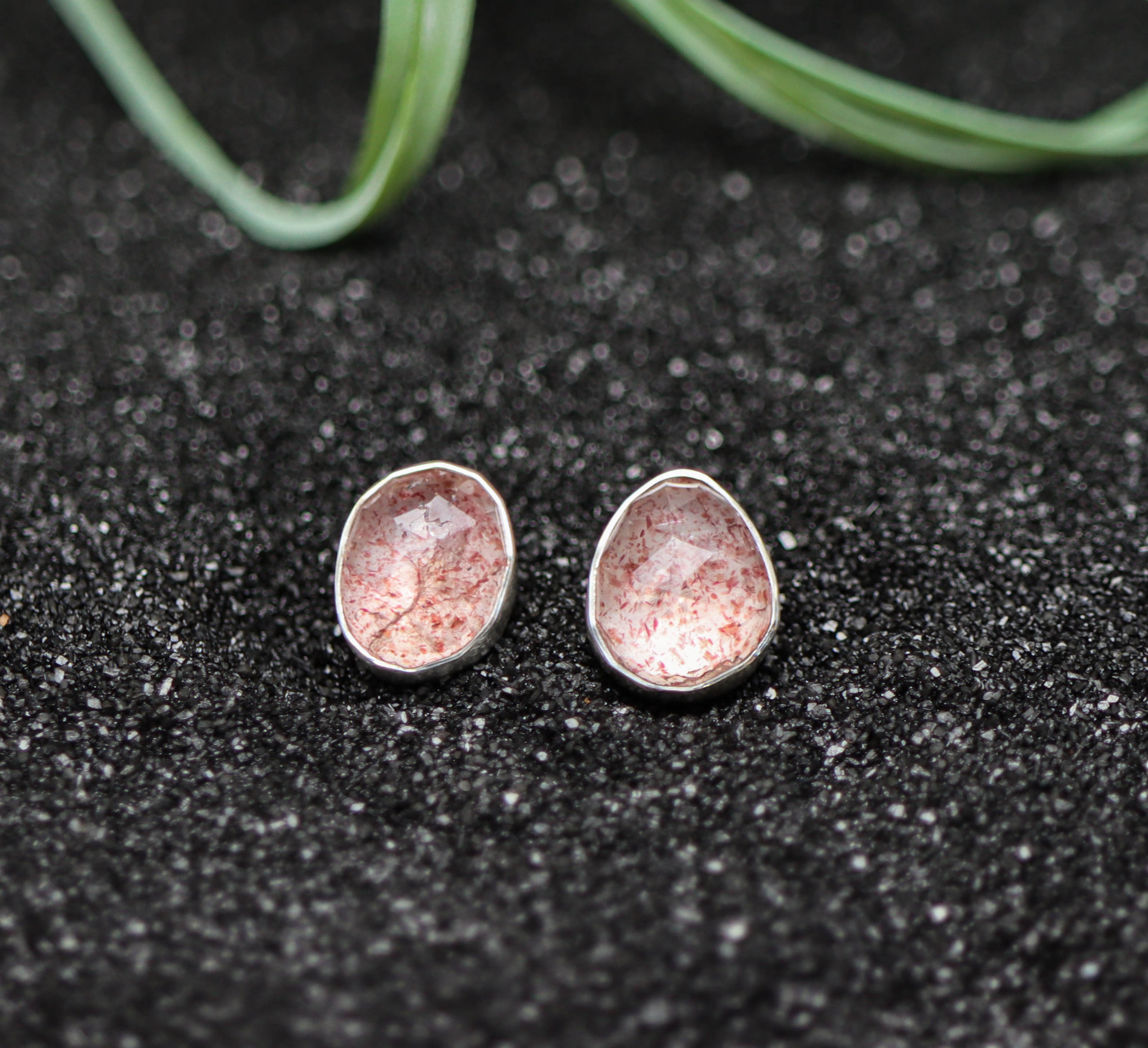Pair of silver earrings with pinkish-red speckles on a dark textured surface.