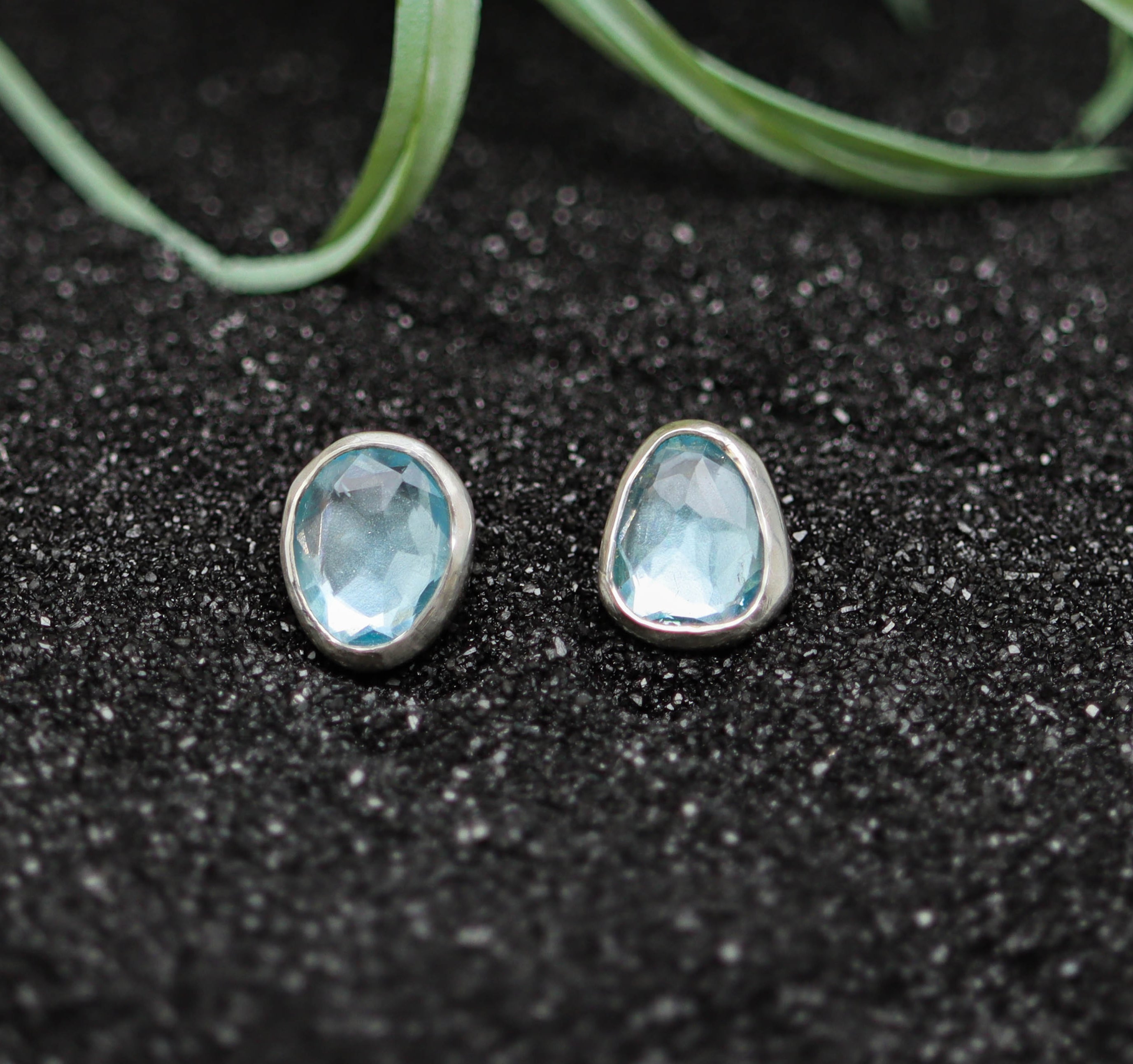 Teardrop-shaped blue gemstone earrings on a black textured surface with greenery in the background.