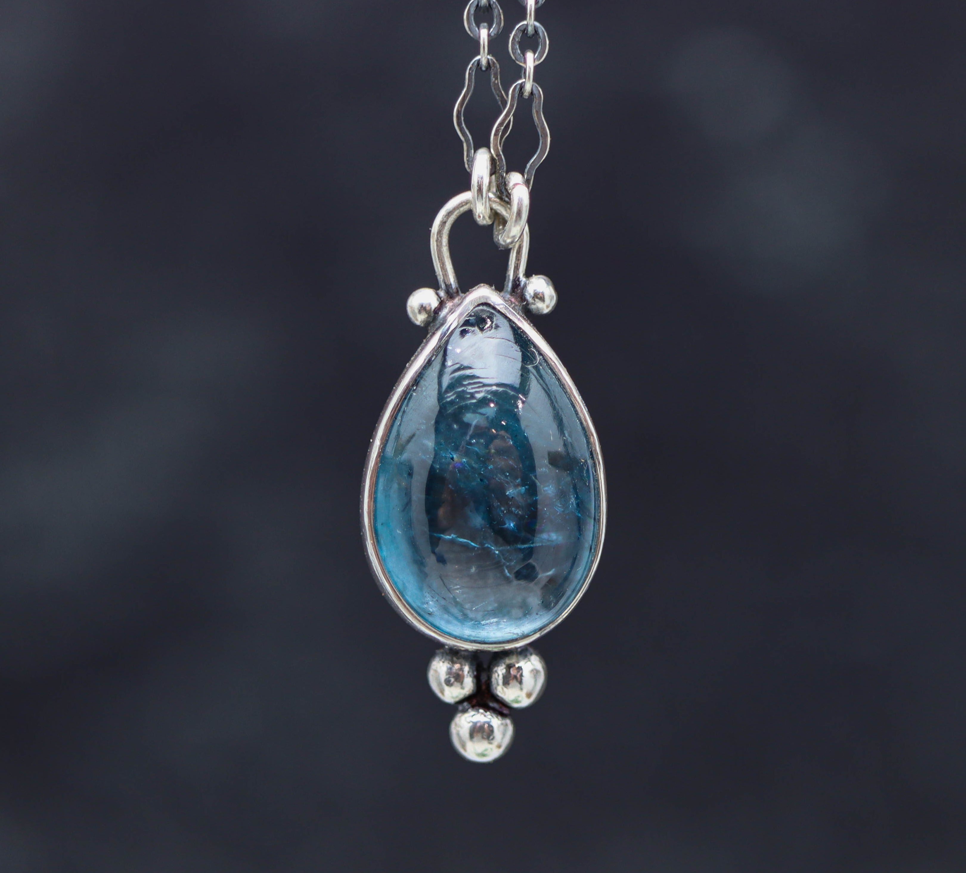 Teardrop-shaped pendant with blue gemstone and silver chain on a dark background