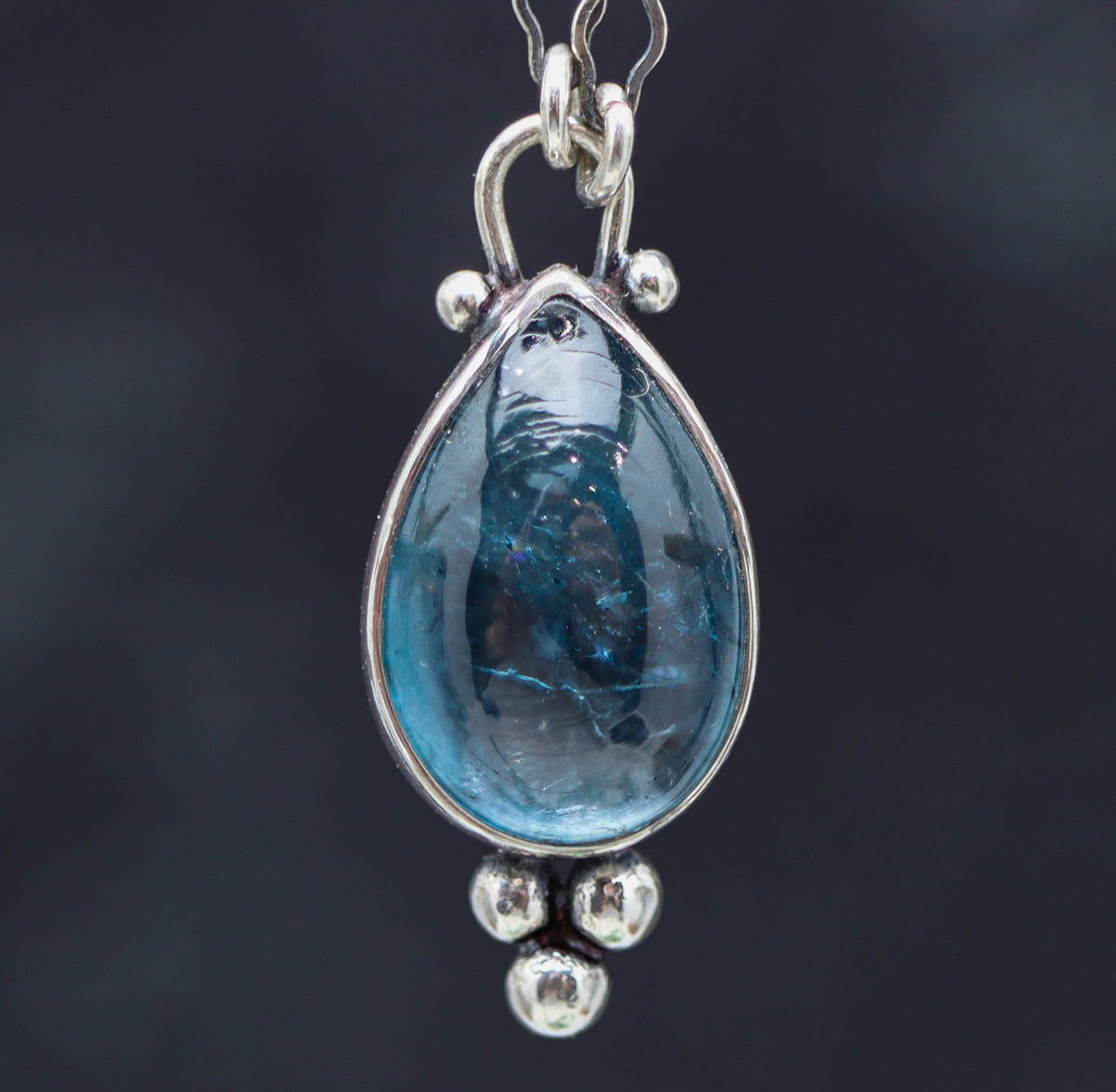 Teardrop-shaped pendant with blue gemstone and silver chain on a dark background